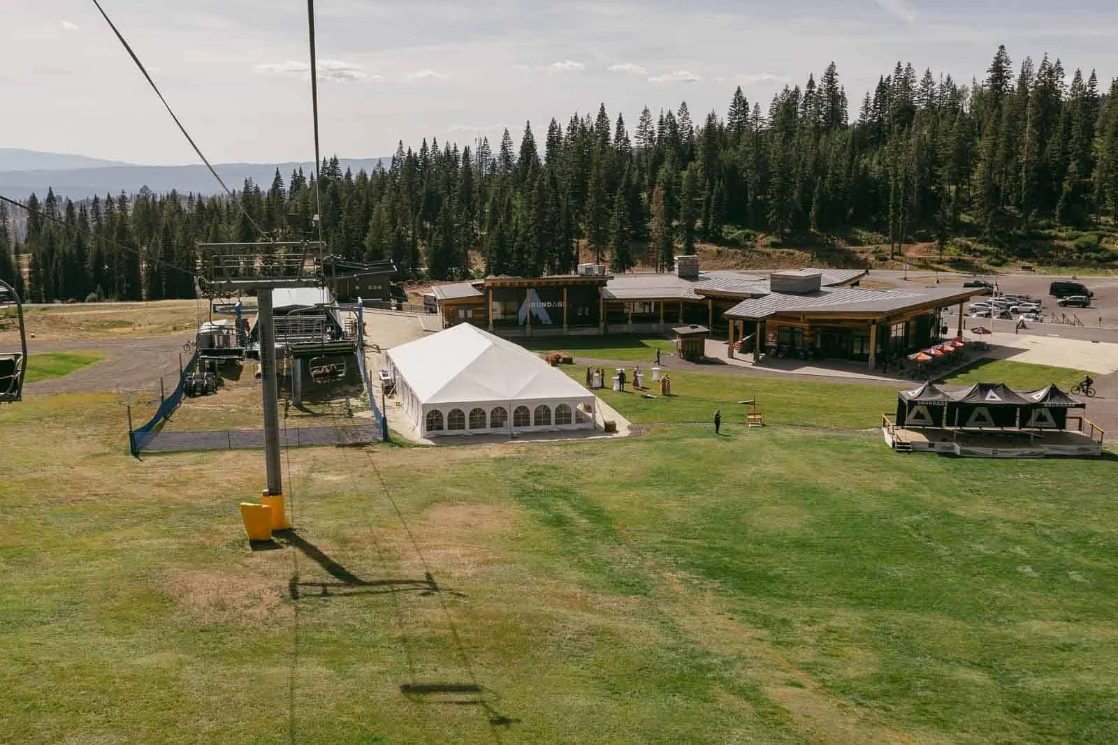 Looking down the Bluebird Chairlift line past empty chairs toward the Brundage Mountain base area, with a white reception tent, lodge buildings, and parking surrounded by pine forest.