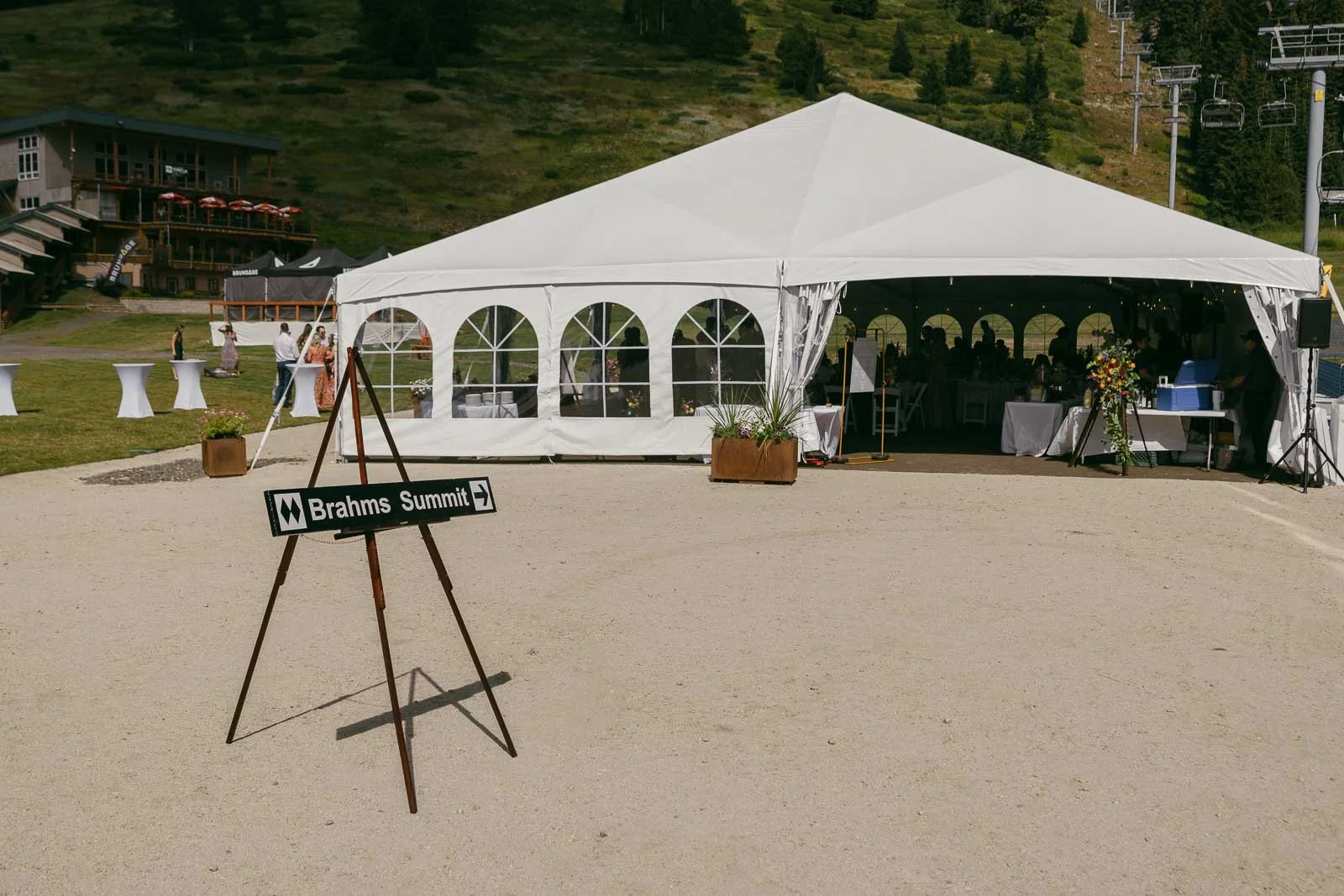 A vintage Brahms Summit ski trail sign on a wooden easel points wedding guests toward the white Bluebird Pavilion reception tent, with flowers, cocktail tables, and chairlift towers on the slope behind.