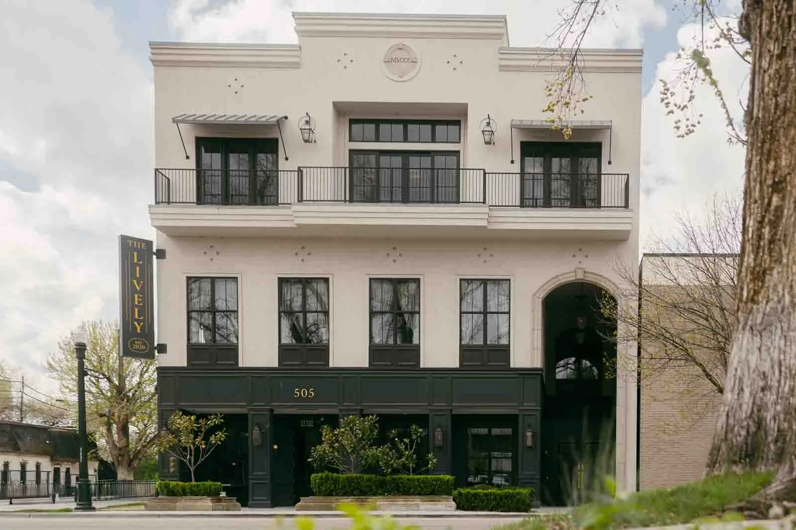 A three-story building with large windows and balconies, a sign reading "The Lively" on the side, and the address "505" above the entrance. Trees and greenery line the sidewalk in front.
