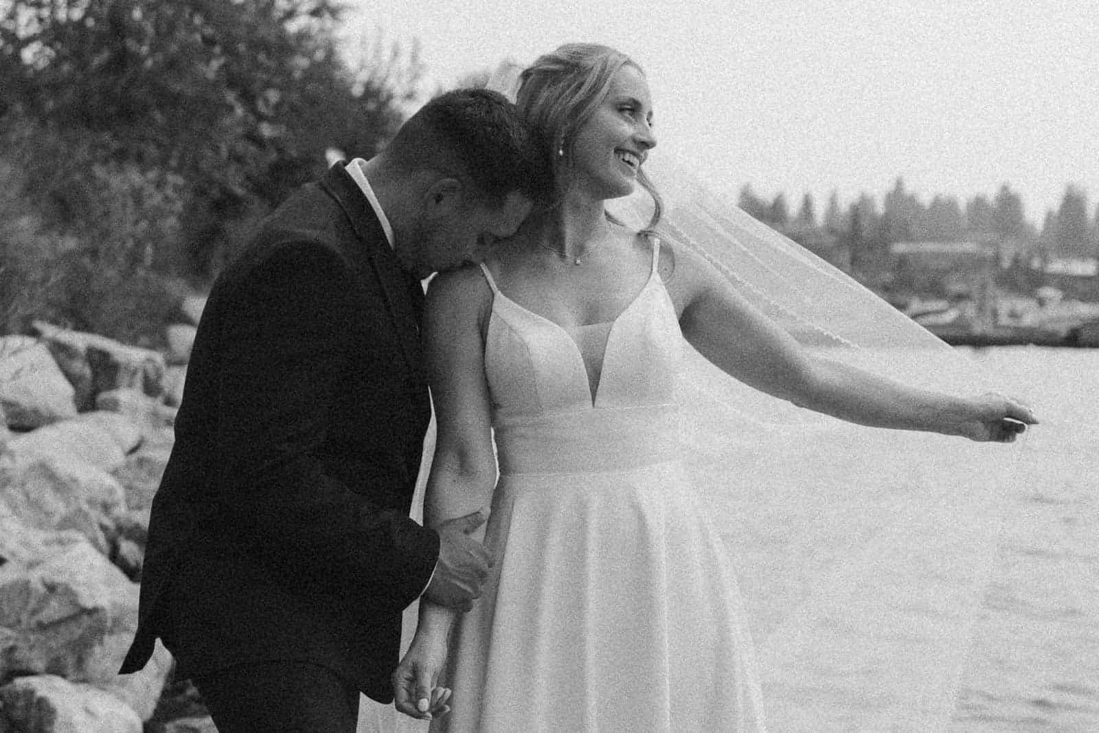Groom tenderly kisses his joyful bride’s shoulder as her veil lifts in the breeze along a McCall, Idaho lake at their wedding