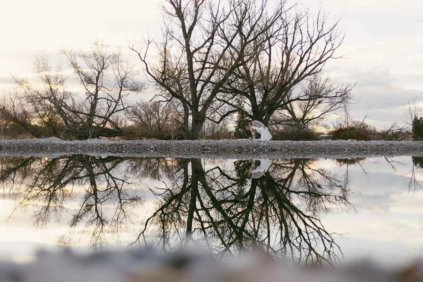 A married couple in white kiss near leafless trees by a calm body of water, with the trees and person reflected clearly in the water under a cloudy sky.