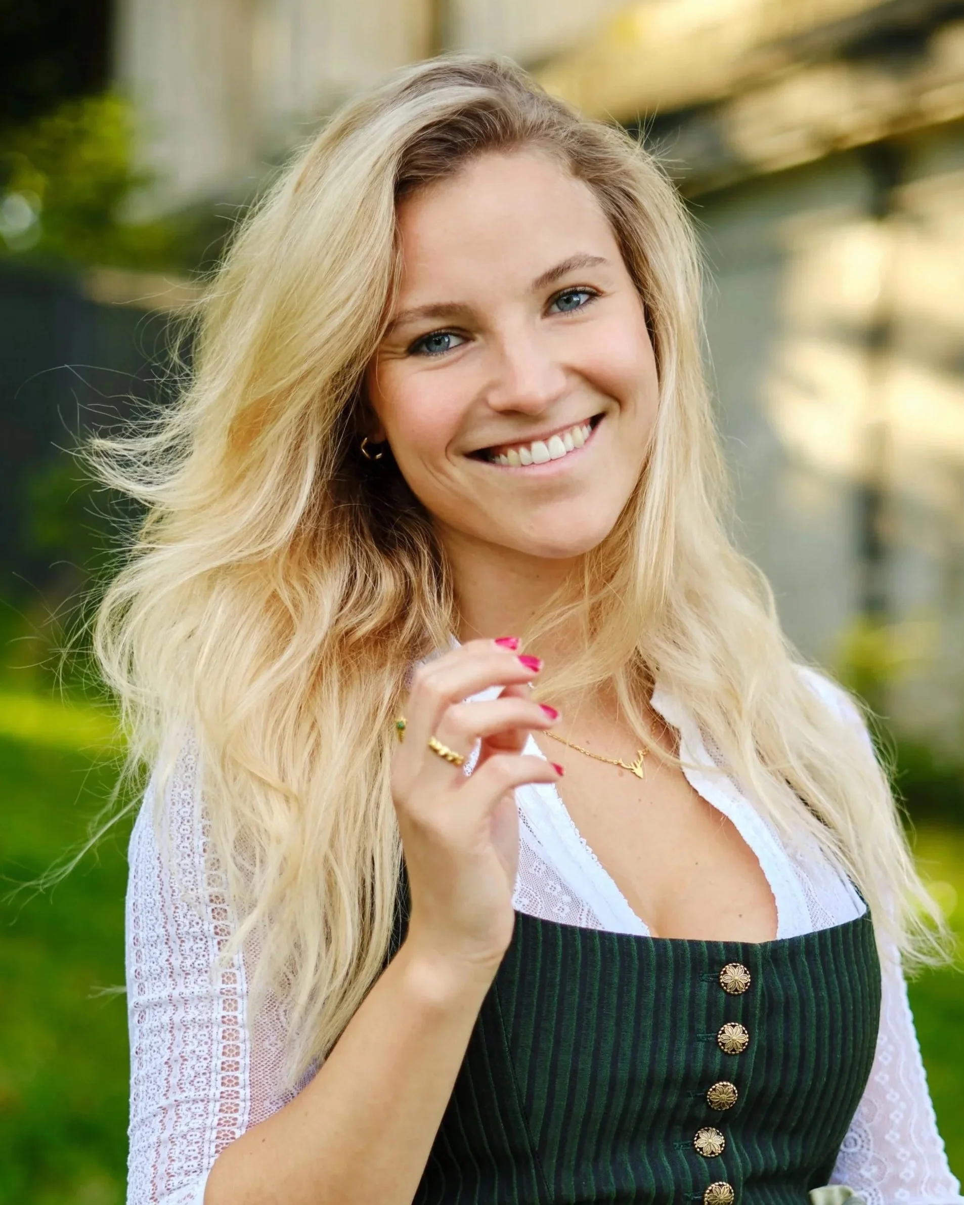 Porträt einer blonden Frau im Dirndl, die lächelnd in die Kamera blickt, vor der Münchner Bavaria im Hintergrund.