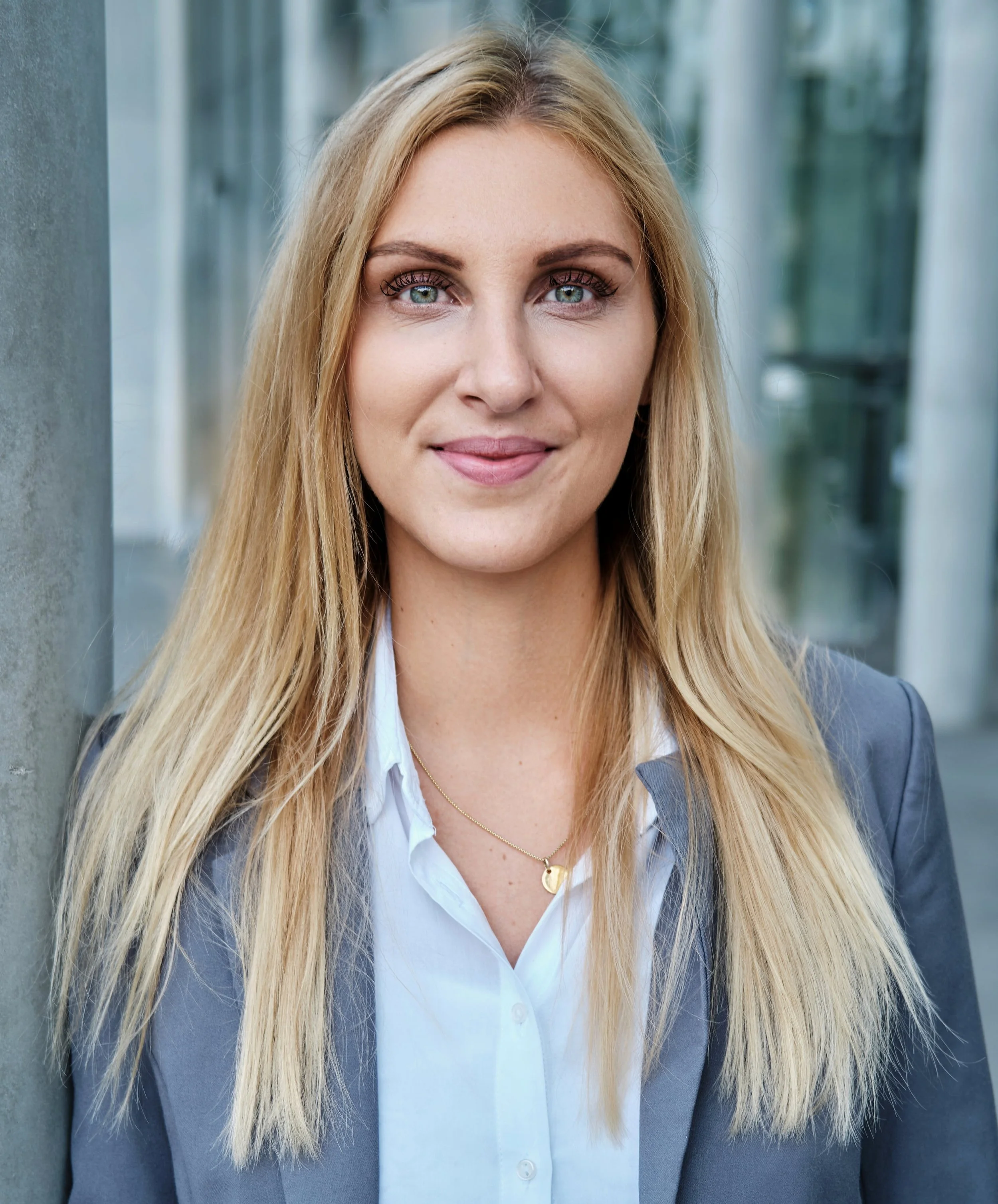 Portrait einer blonden jungen Frau im Anzug, die mit leicht neutralem Lächeln in die Kamera blickt, vor einem grünen und weißen Hintergrund, in dem Fenster und Säulen zu sehen sind.
