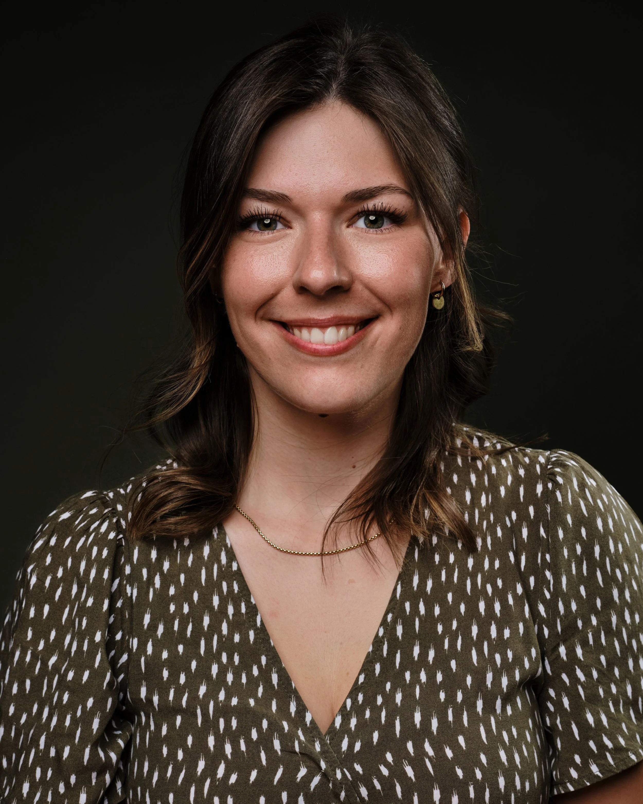 Porträt einer blonden jungen Frau, die lächelnd in die Kamera blickt, vor einem grünem Hintergrund.