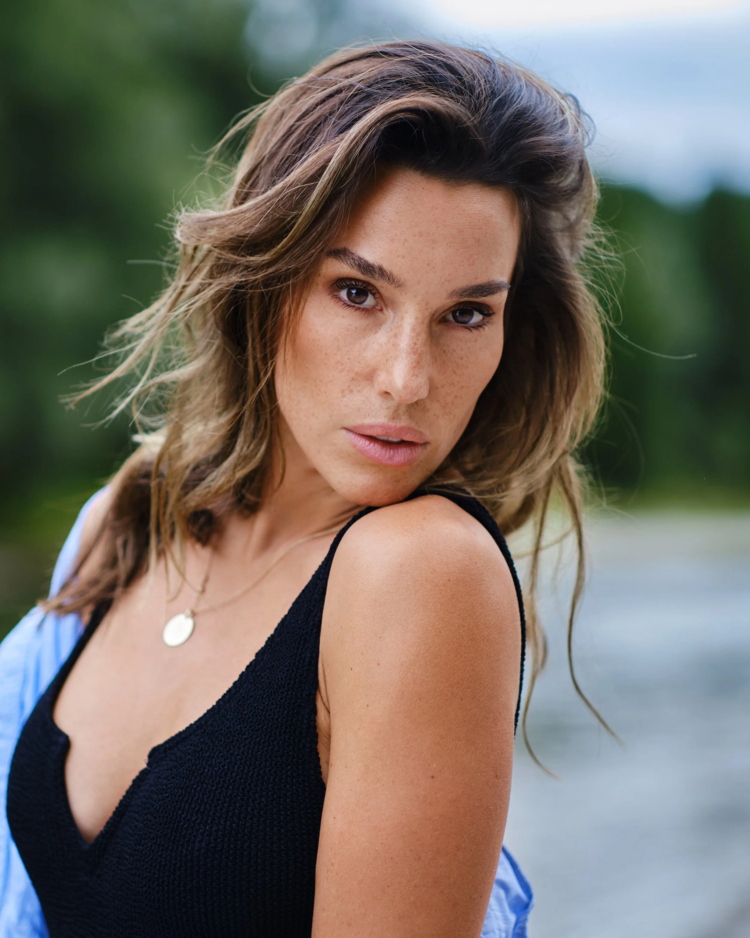 Porträt einer brünetten Frau, die intensiv in die Kamera blickt, vor einem Fluss mit Bäumen im Hintergrund.