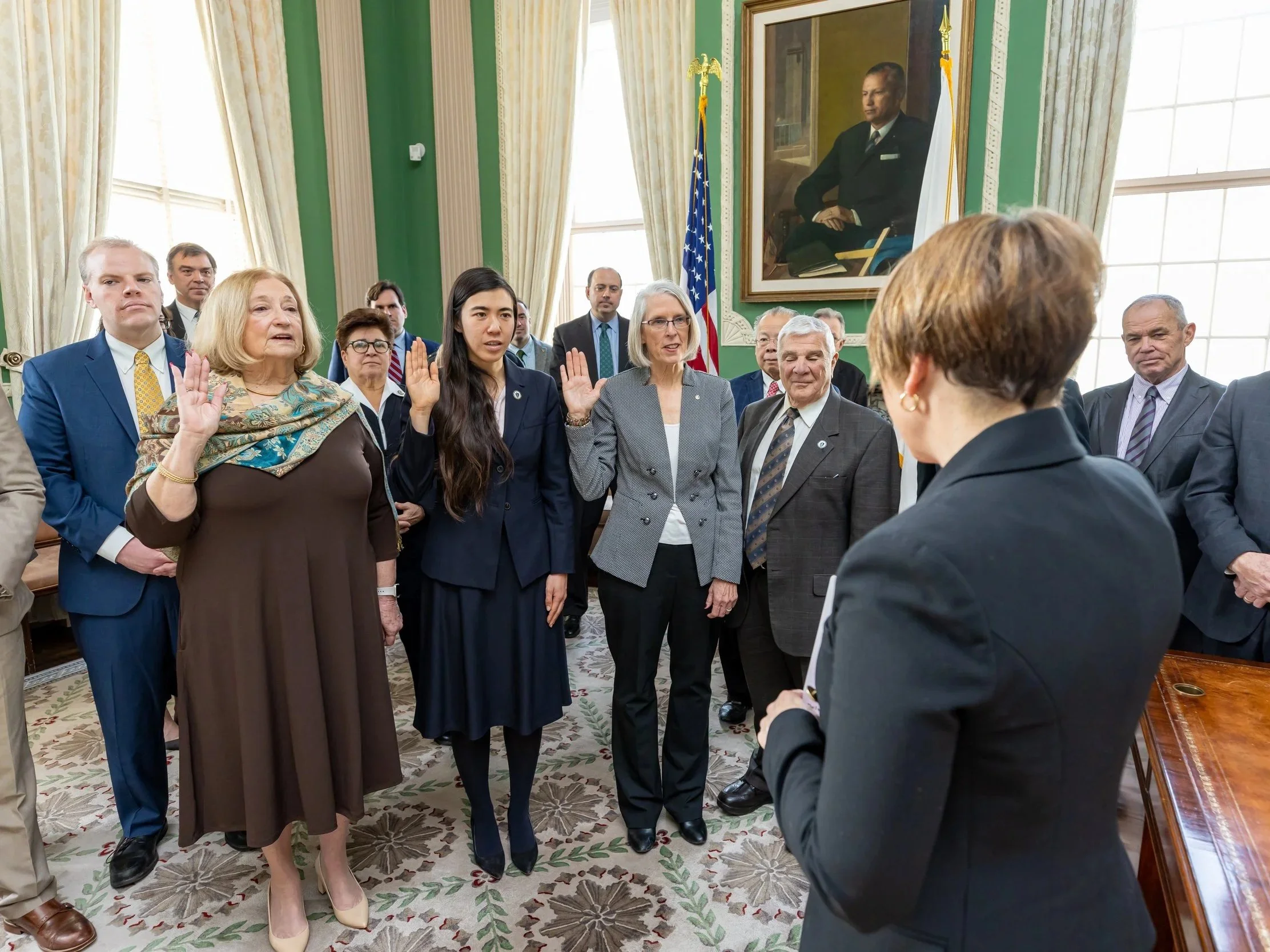 A group of people participating in an oath-taking ceremony in a formal room with green walls, large windows, and a portrait of a man in military uniform hanging on the wall. Some individuals have their right hands raised, and a woman is facing the group.