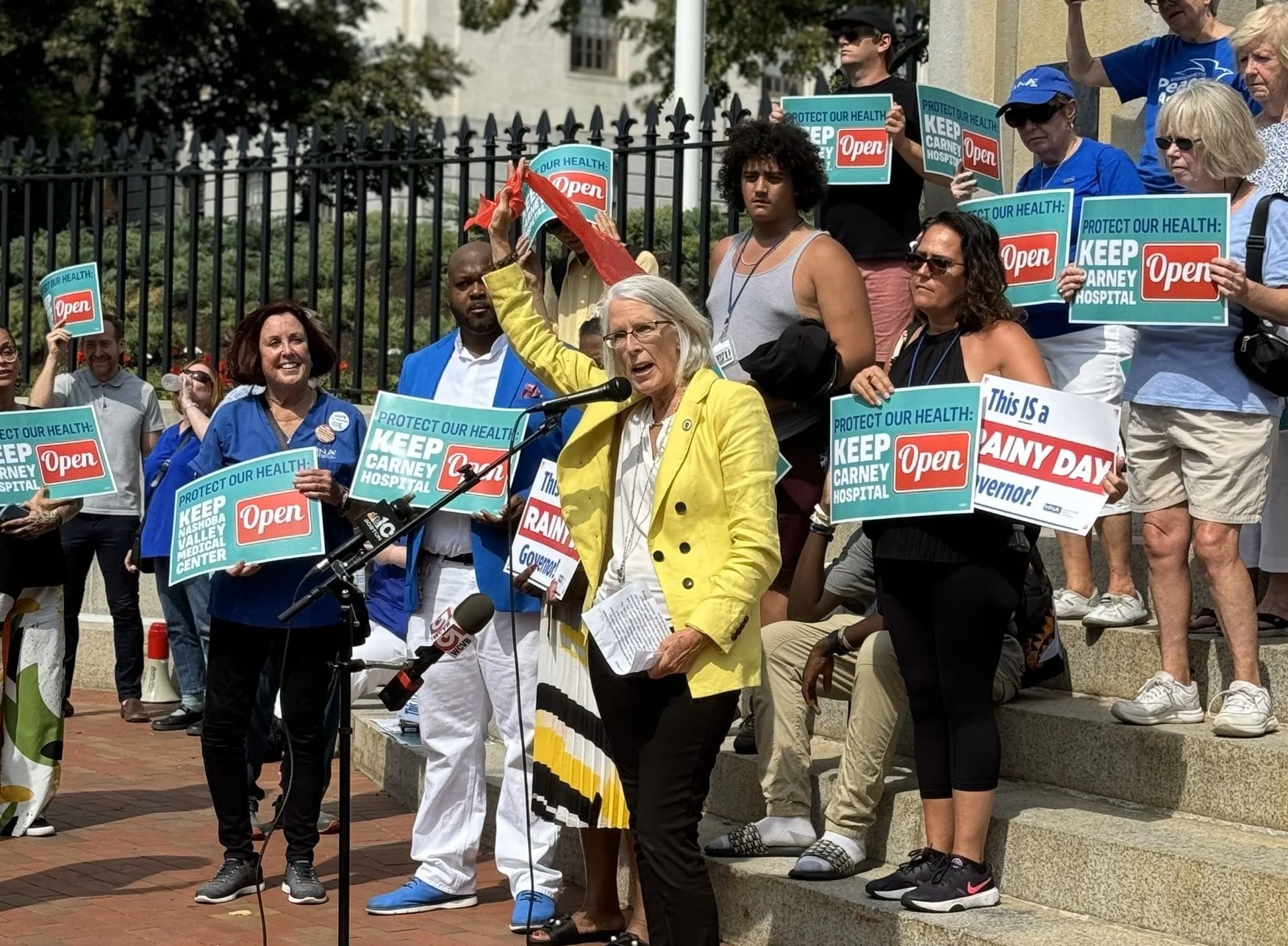 Group of people at a protest holding signs that say 'Protect Our Health: Keep Carney Hospital Open' and 'This is a Dainy Day.' A woman in a yellow jacket is speaking at a microphone. Several individuals are wearing sunglasses and casual clothing, and some are standing on steps.