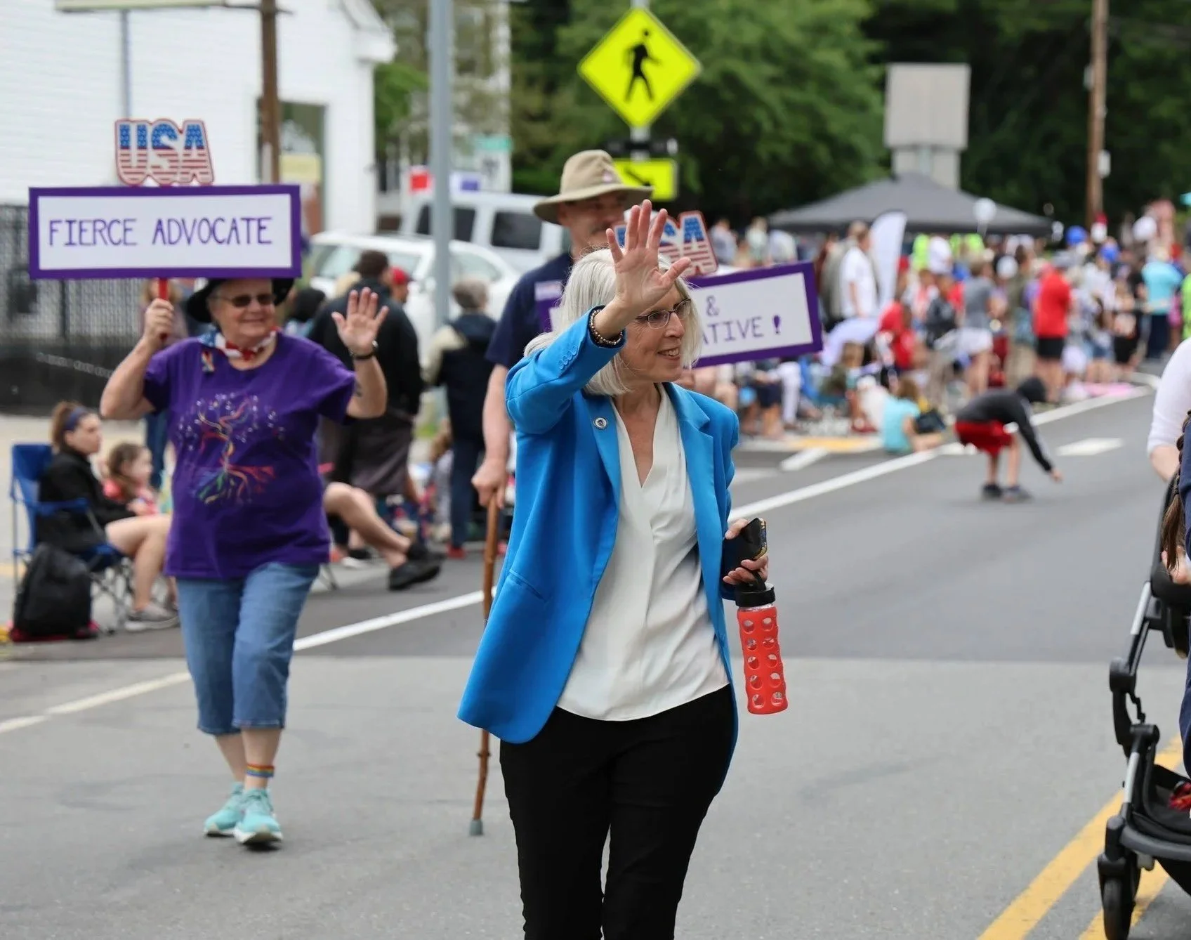 A group of people participating in a parade or rally on a street, with some holding signs. The signs read 'FIERCE ADVOCATE' and others. People are waving, talking, and some are sitting on the sidewalk. There are vehicles, trees, and a yellow pedestrian crossing sign in the background.