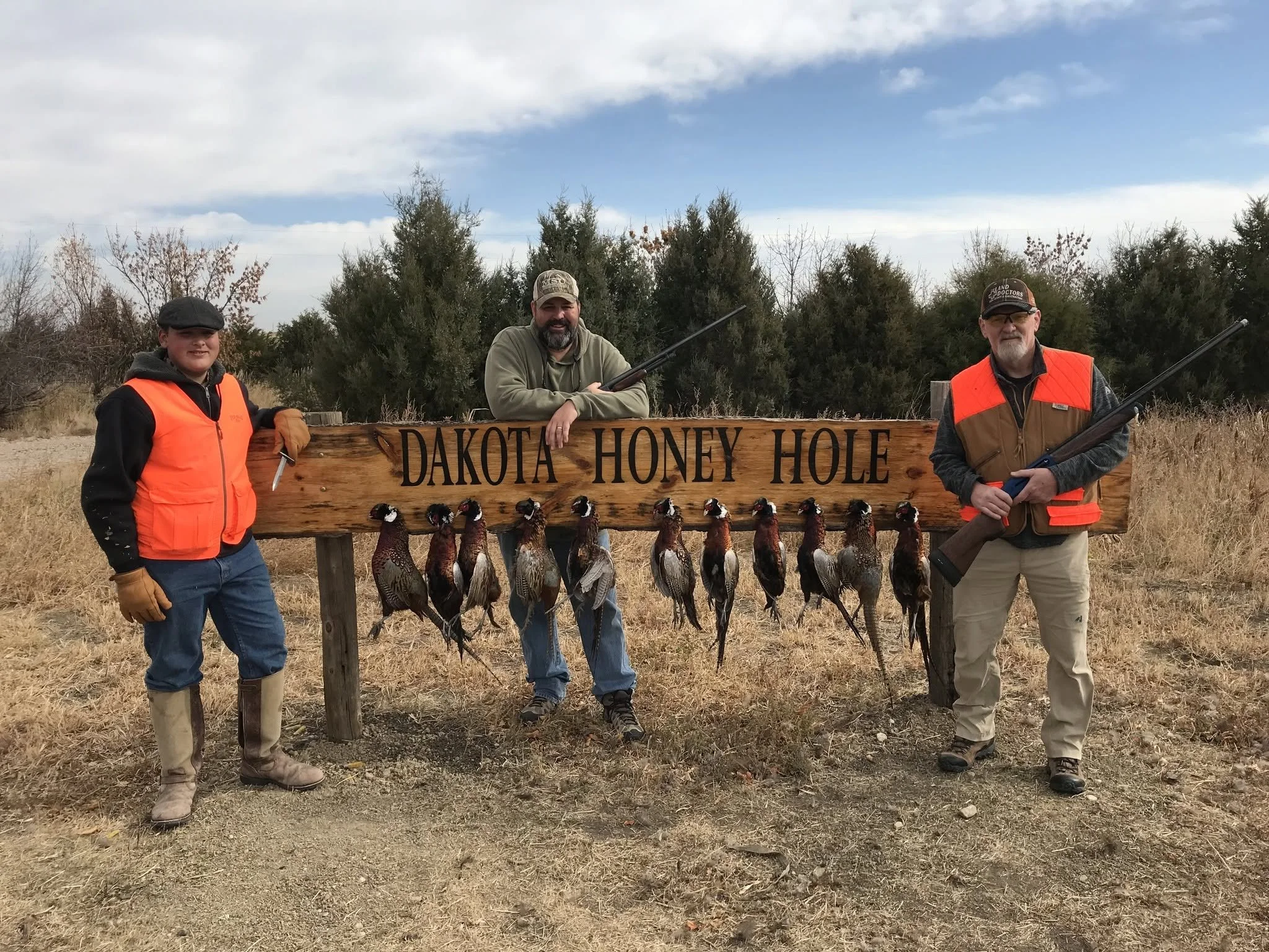 Three people standing outdoors in front of a wooden sign that reads 'Dakota Honey Hole' with dead game birds hanging from it, holding rifles, and wearing hunting gear, with trees and a partly cloudy sky in the background.