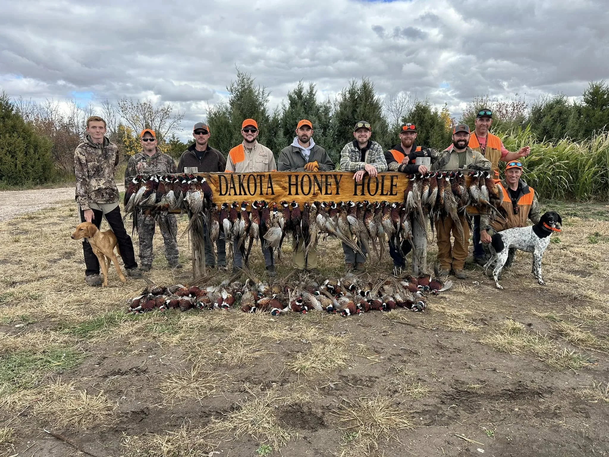 Group of hunters standing behind a wooden sign reading "Dakota Honey Hole" with a variety of ducks hung in front of it, outdoors on a cloudy day.