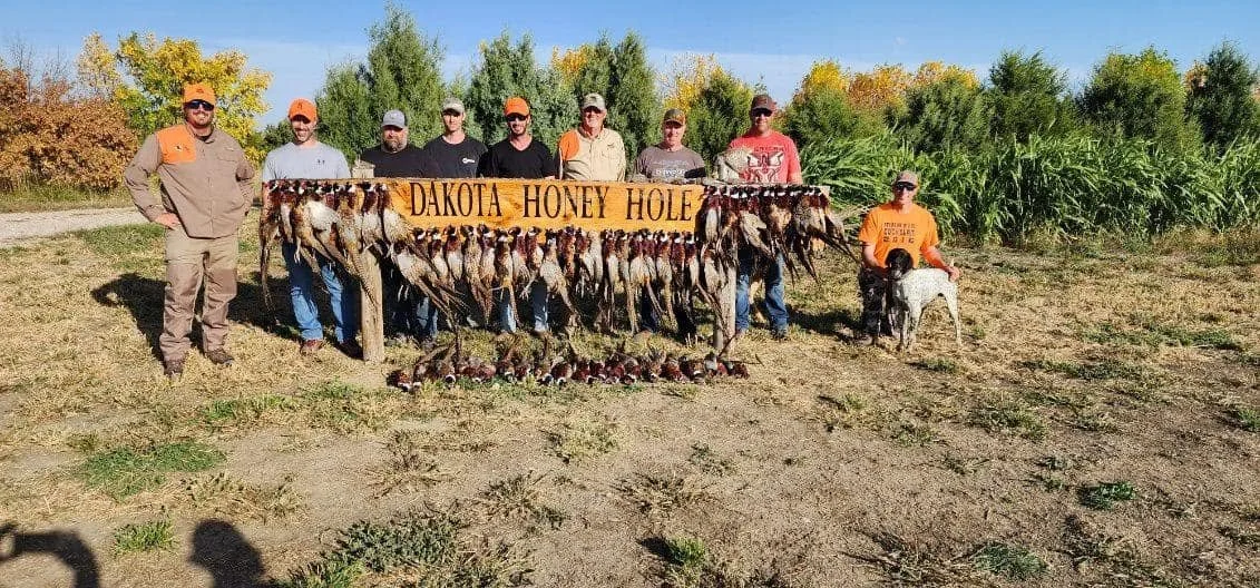 Group of people standing behind a display of harvested ducks and pheasants, with a sign that reads 'Dakota Honey Hole,' in an outdoor field with trees and blue sky.