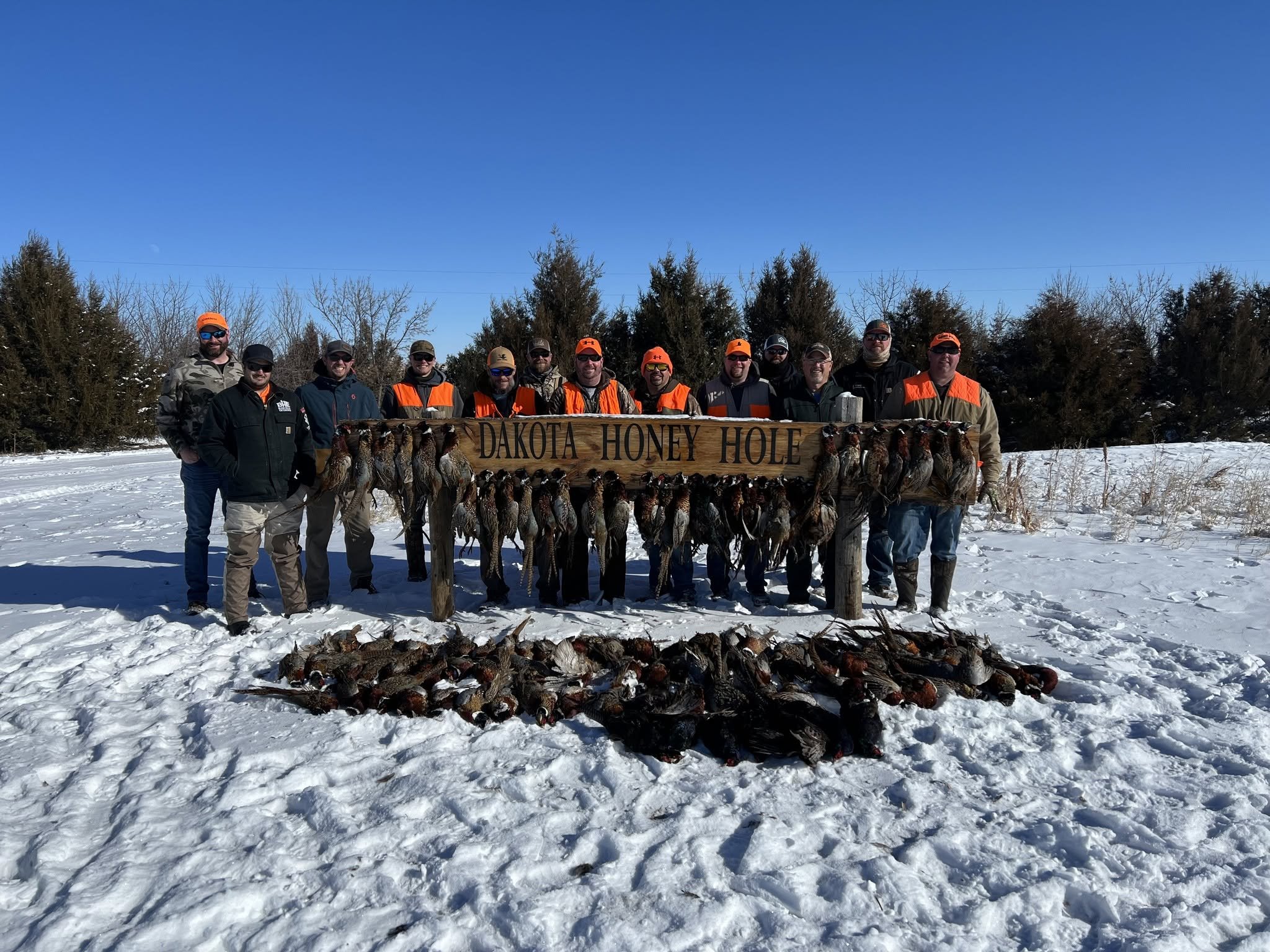 Group of men in outdoor clothing standing behind a wooden sign reading "Dakota Honey Hole" with a row of ducks hung on the sign in a snowy landscape under a clear blue sky.
