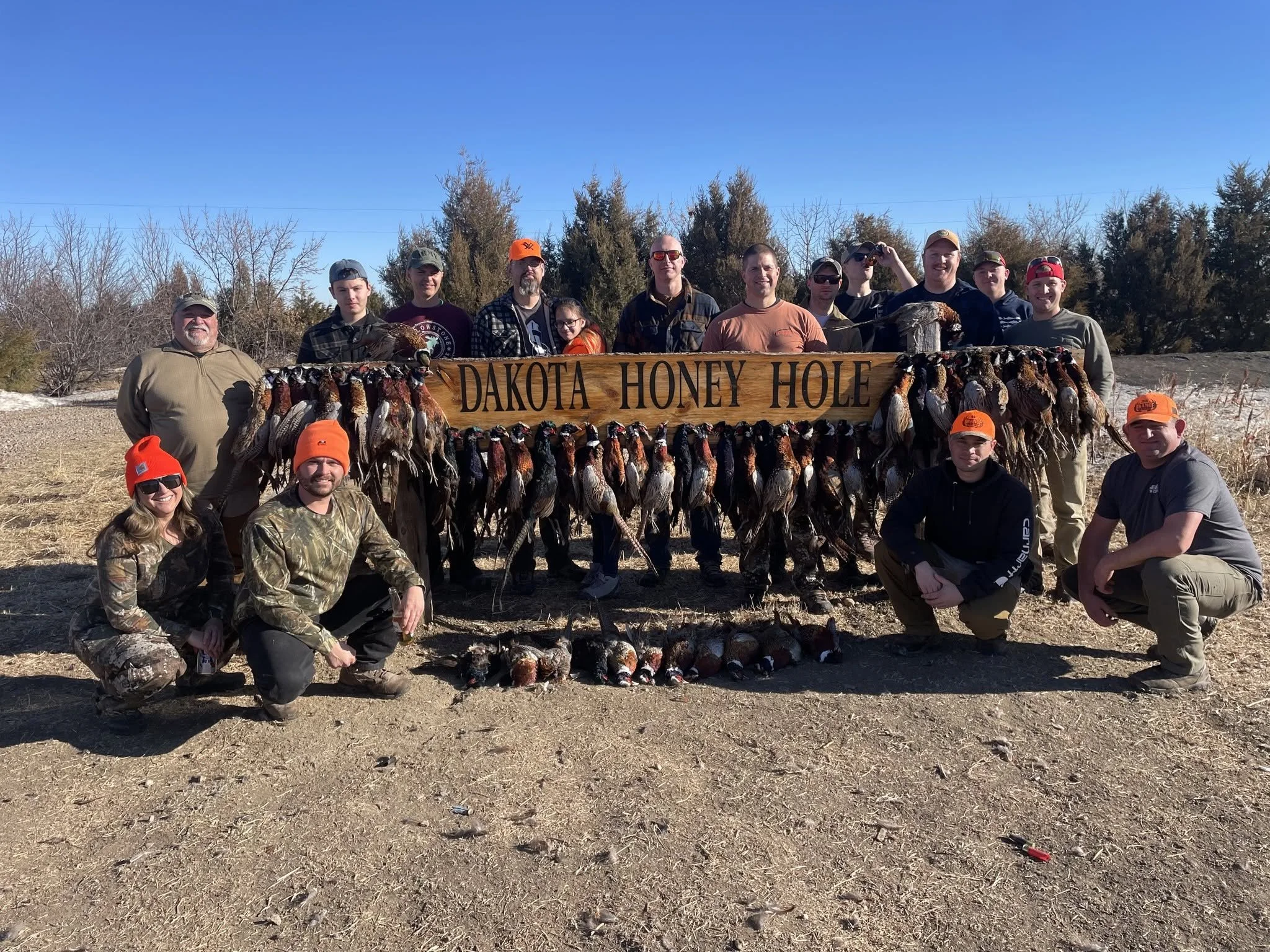 Group of people outdoors holding a large wooden sign that reads 'Dakota Hony Hole', with numerous harvested ducks hanging from the sign. The group is dressed in outdoor and camouflage clothing, smiling, and standing under a clear blue sky with trees 