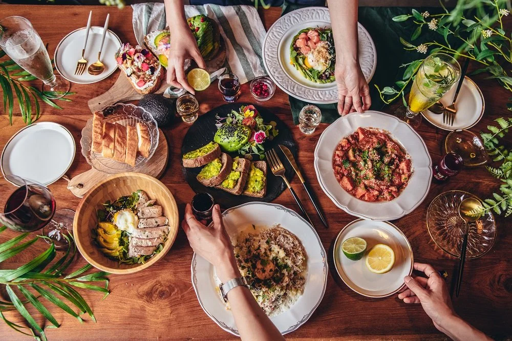 private chef dinner table with diverse dishes, drinks, and people reaching for food, surrounded by plants and cutlery.