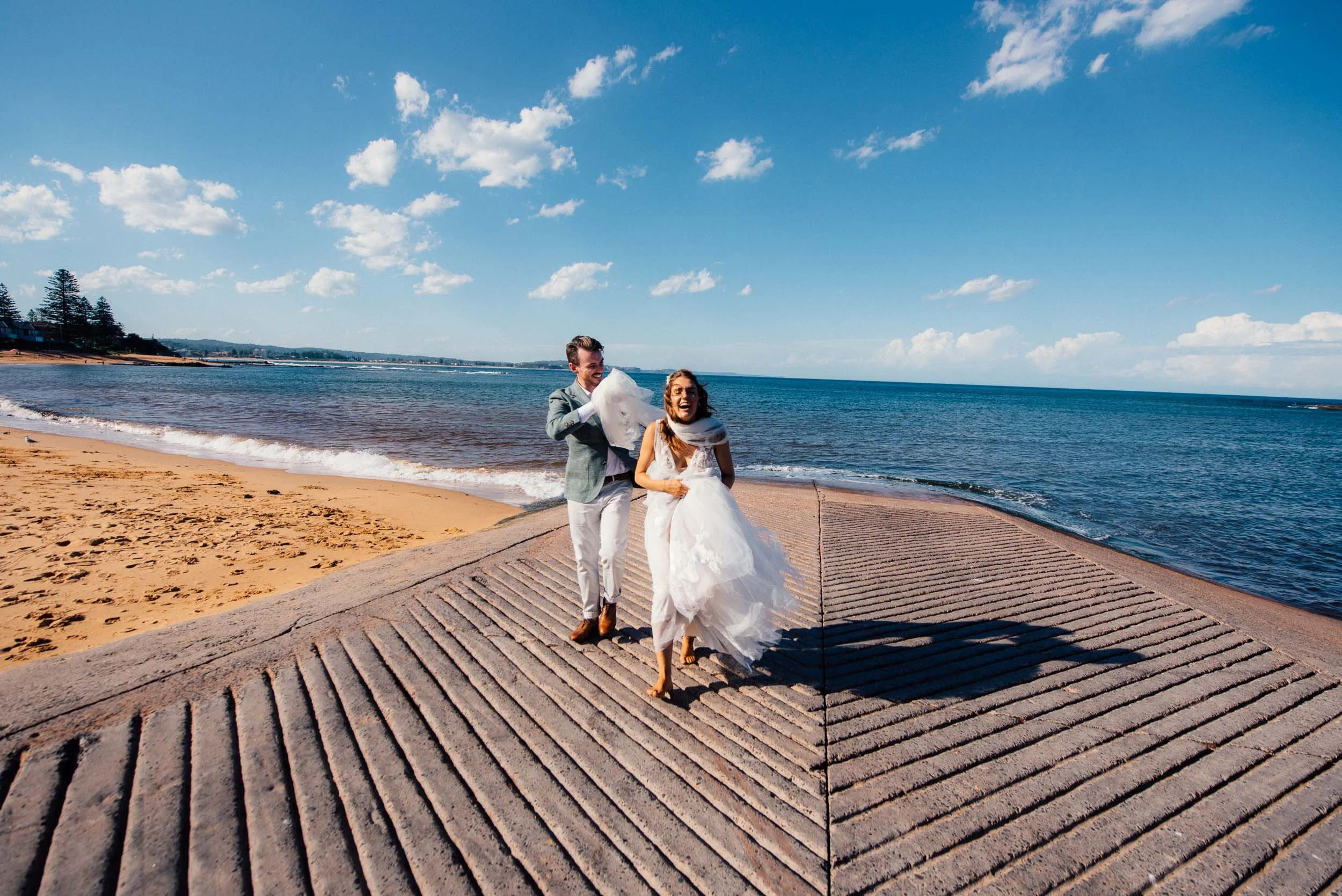 A bride and groom in wedding attire running along a seaside walkway on a sunny day, with the ocean and blue sky in the background.