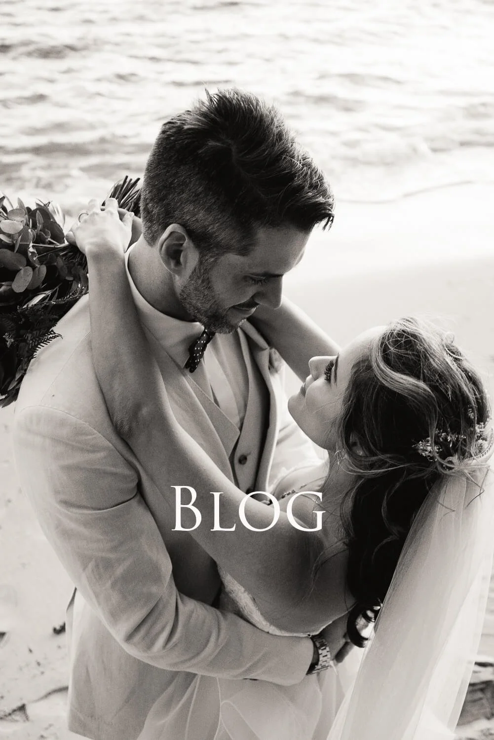 A black and white photo of a groom and bride embracing on the beach, gazing into each other's eyes. The groom holds a bouquet, and the bride has a floral hairpiece. The word "BLOG" is overlaid on the image.