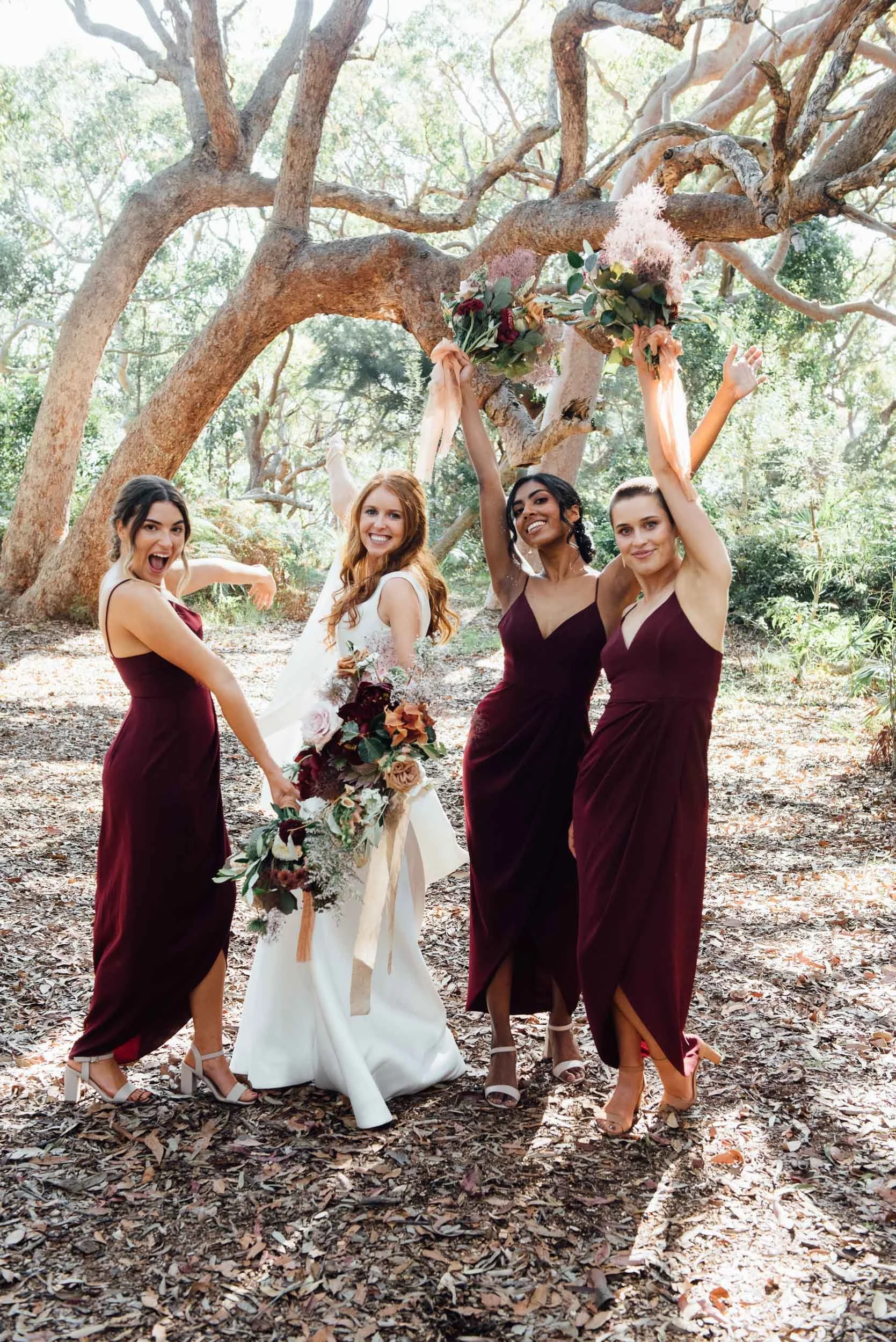 Four women, including a bride in a white dress, and three bridesmaids in burgundy dresses, posing outdoors in a wooded area, holding and showcasing floral arrangements and celebrating.