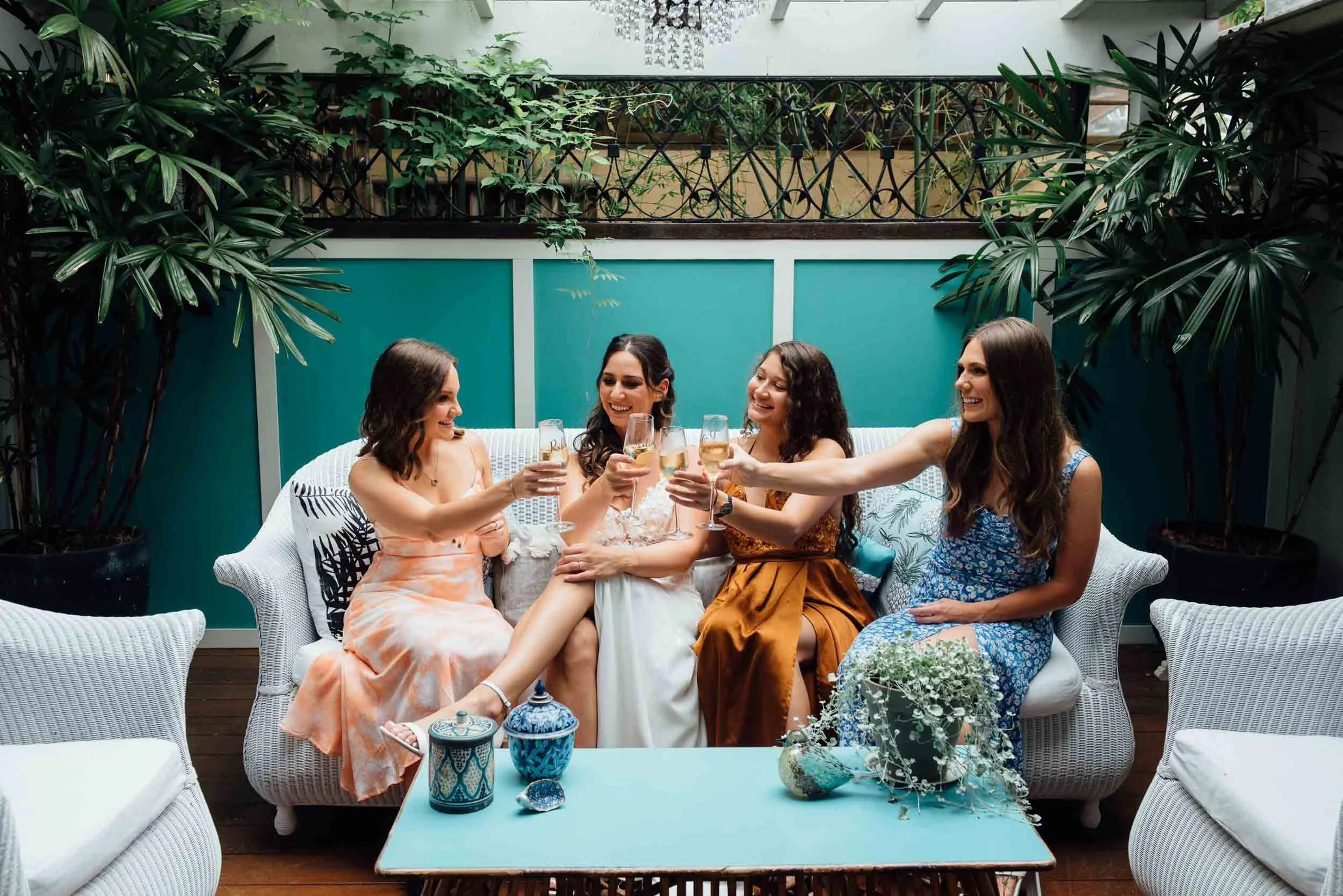 Four women celebrating with champagne in a cozy indoor patio with teal and white decor, surrounded by greenery.