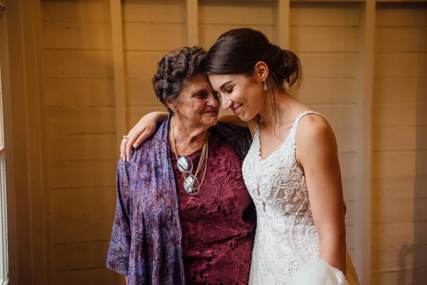A bride and an elderly woman sharing a tender moment with foreheads touching and eyes closed, in a warmly lit room with wood-paneled walls.