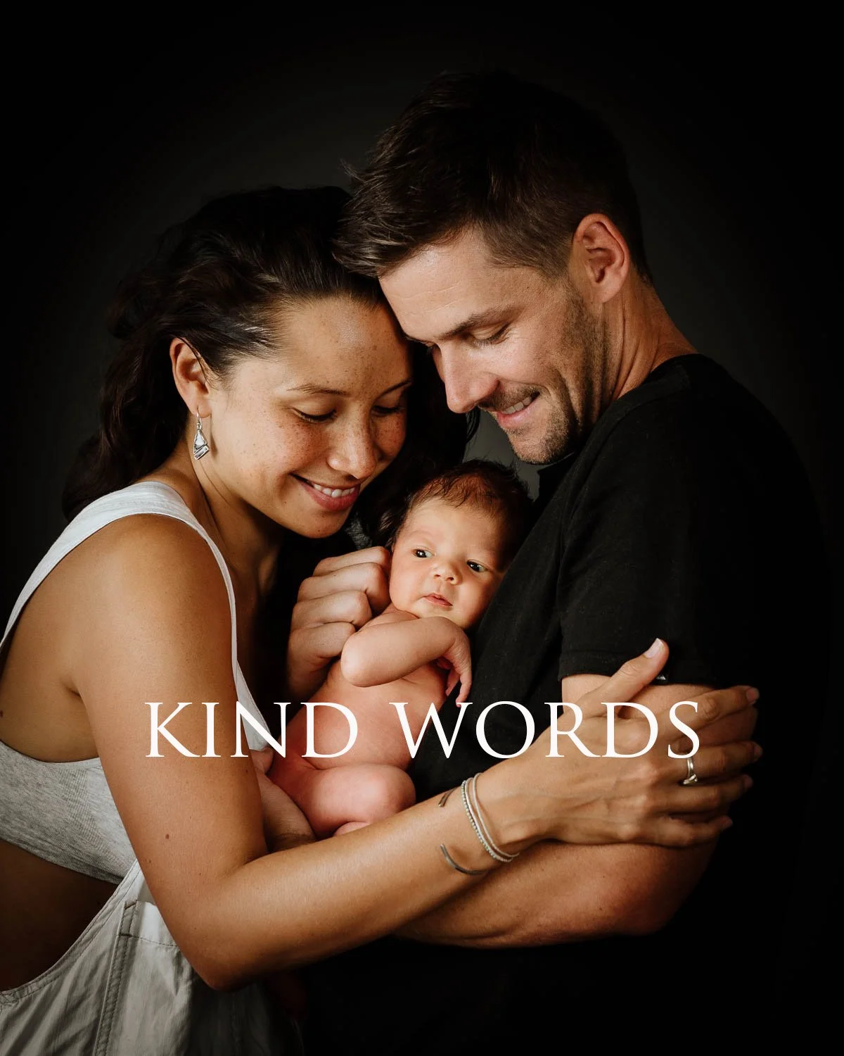 A happy family holding a newborn, with a woman and man smiling and the baby looking curious. The words 'KIND WORDS' are overlaid on the image.