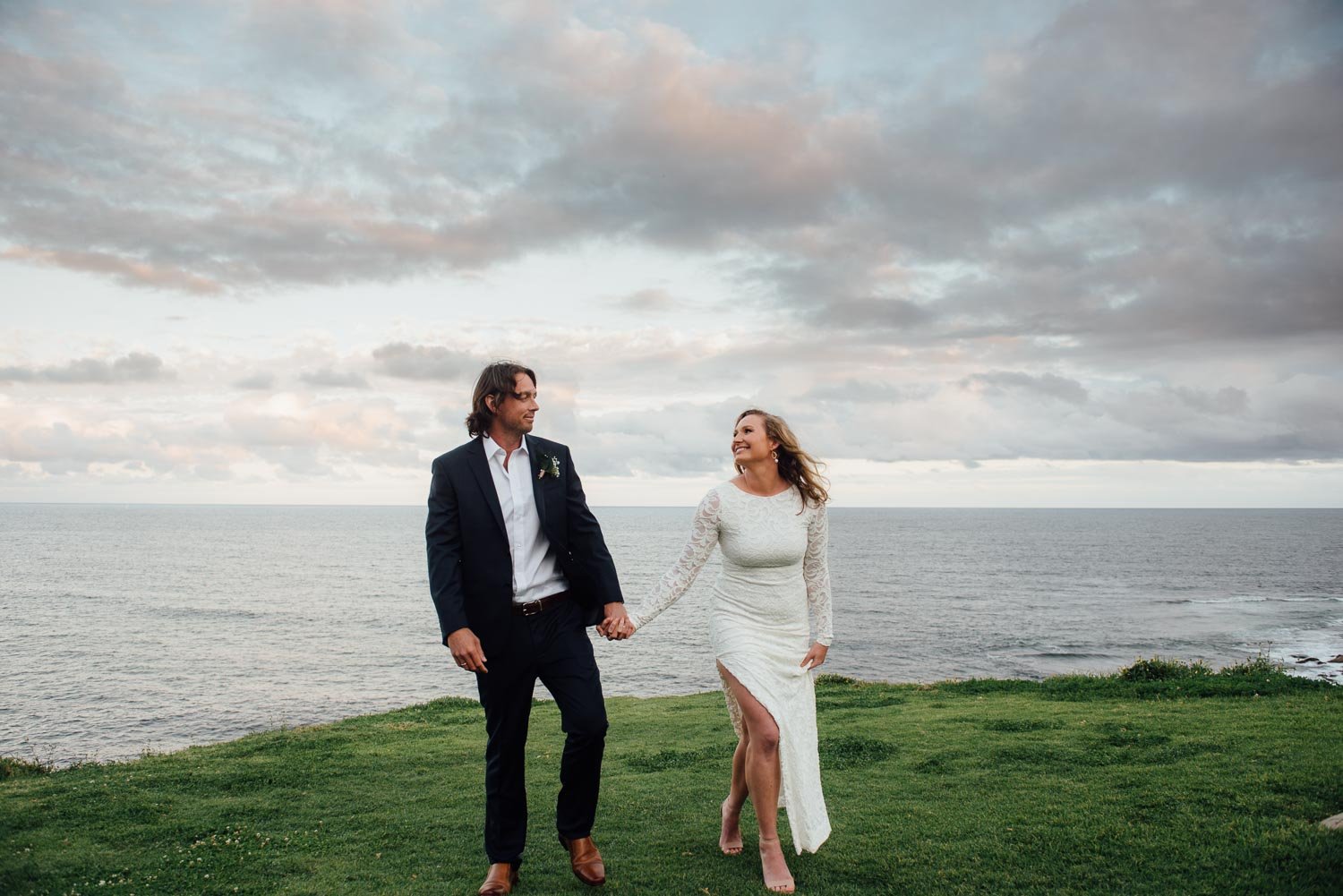 A couple holding hands and walking along a grassy area near the ocean at sunset, with clouds in the sky. The woman wears a white lace dress with a slit, and the man wears a dark suit with a white shirt.