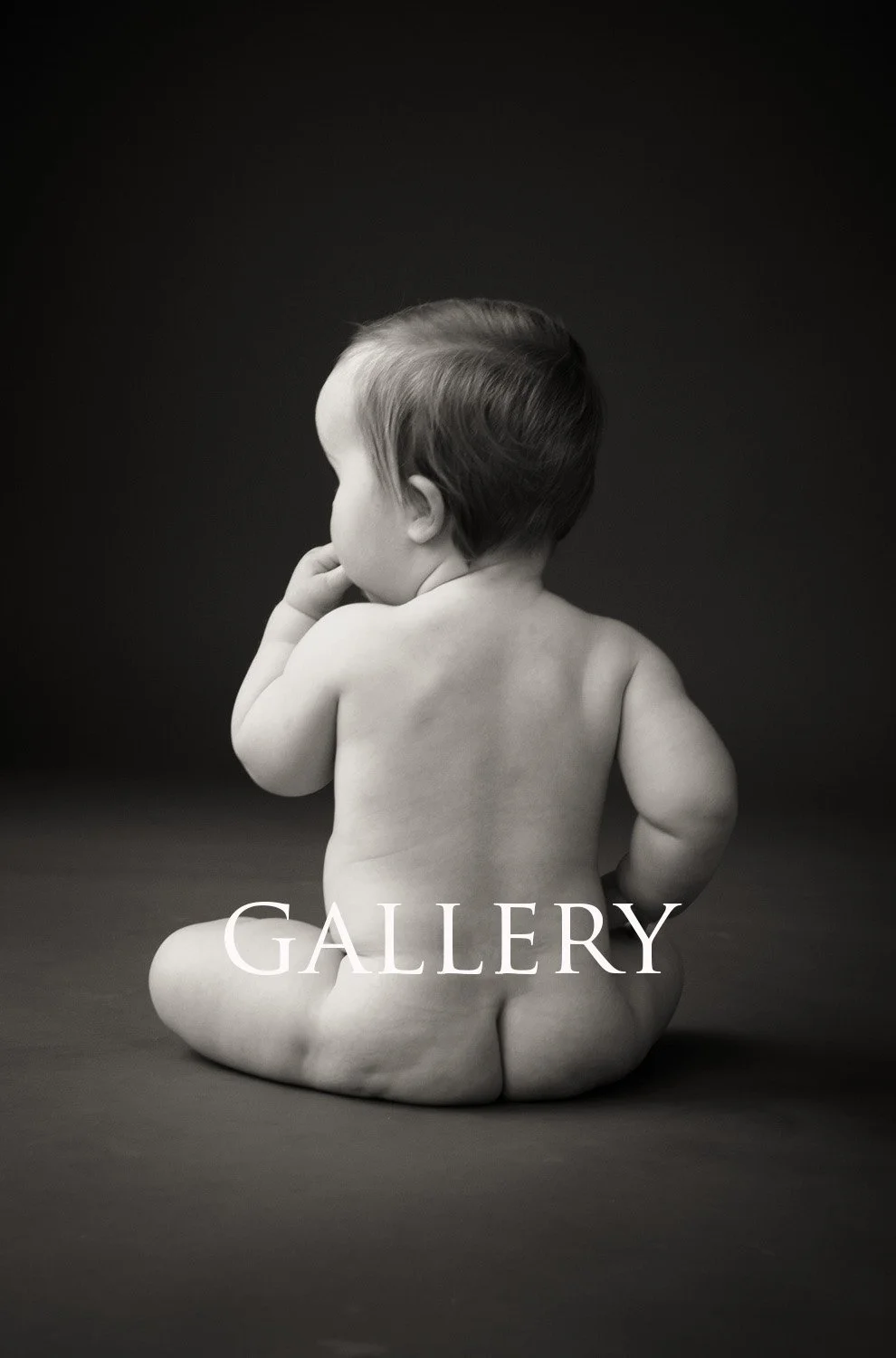 Black and white photograph of a young nude child sitting with their back to the camera against a dark background, with the word 'GALLERY' overlaid.