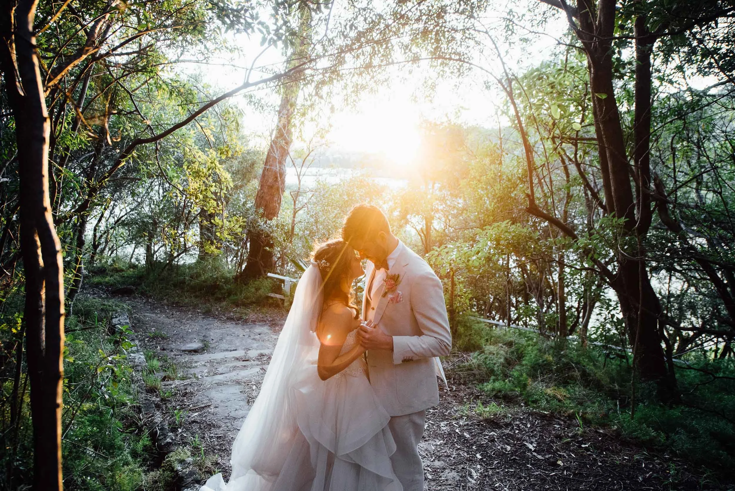 A bride and groom in wedding attire dancing closely outdoors at sunset in a wooded area.
