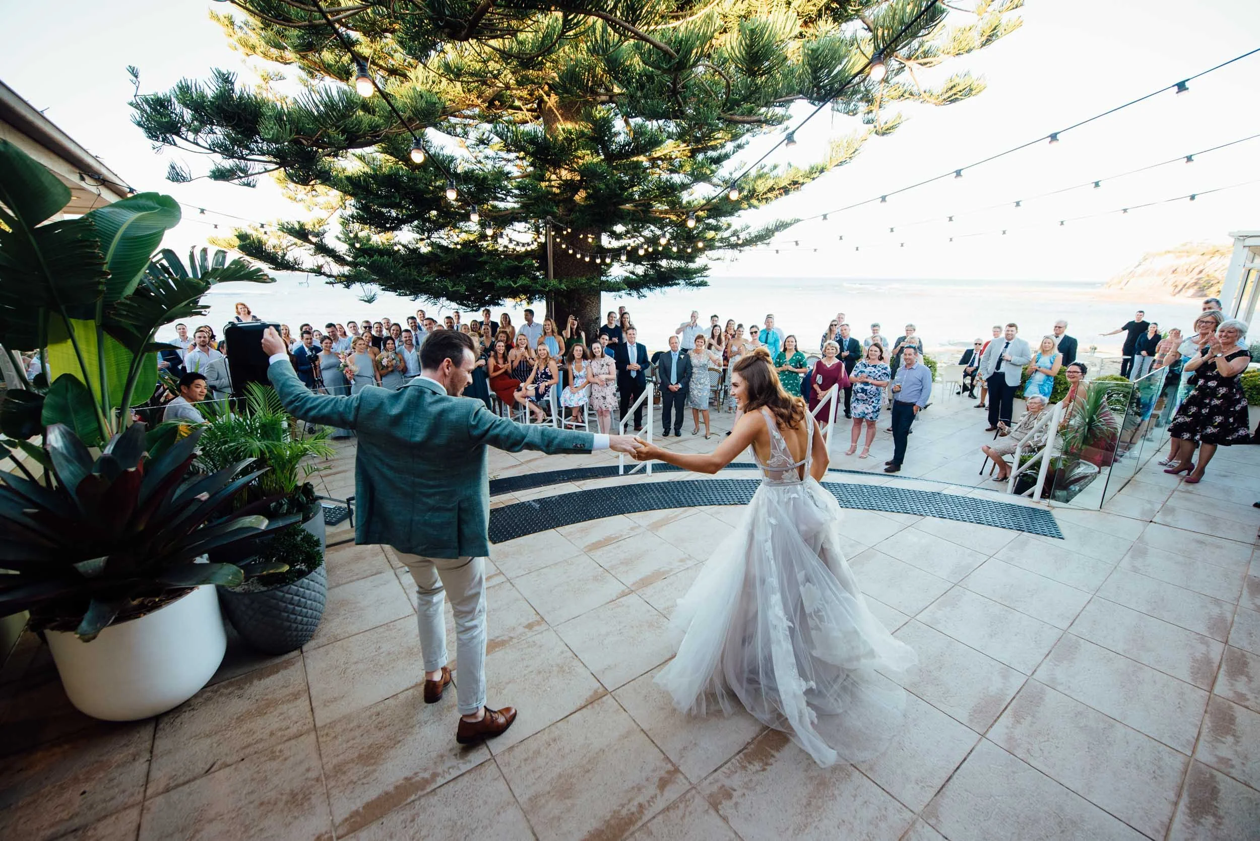 A bride and groom dance at their wedding reception outdoors by the ocean, with guests watching under a large pine tree decorated with string lights.