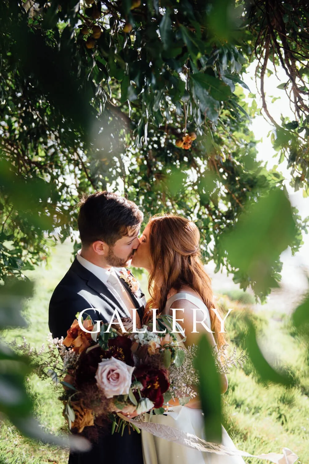 A bride and groom share a kiss outdoors beneath a tree, with sunlight filtering through the leaves. The bride has long red hair and wears a white gown, holding a bouquet of flowers. The groom wears a dark suit and white shirt. The word "GALLERY" is w