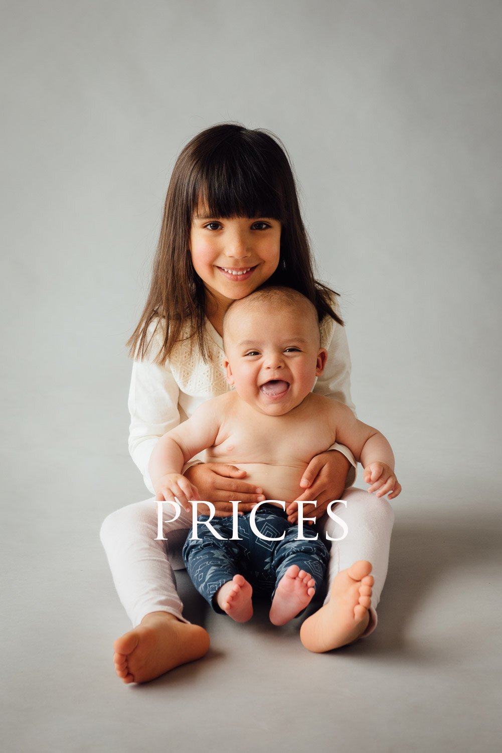 Upset baby sitting on lap of young girl with dark hair and smiling face, against plain gray background.