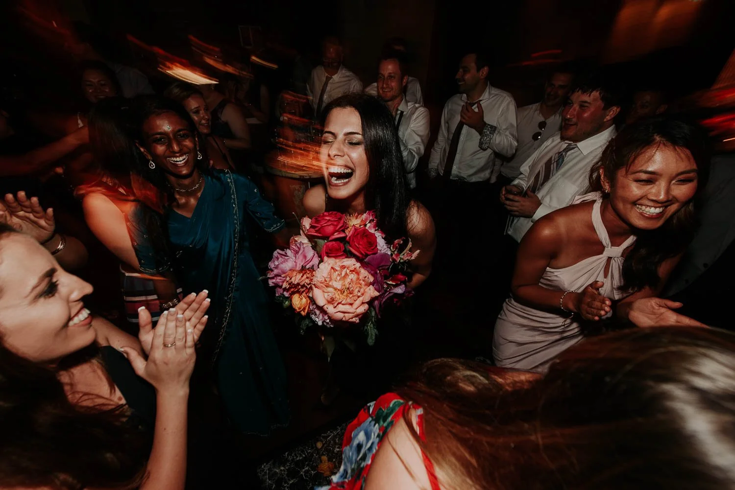 Woman in black dress holding a bouquet of red, pink, and orange flowers, smiling and crying tears of joy, surrounded by friends celebrating at a party.