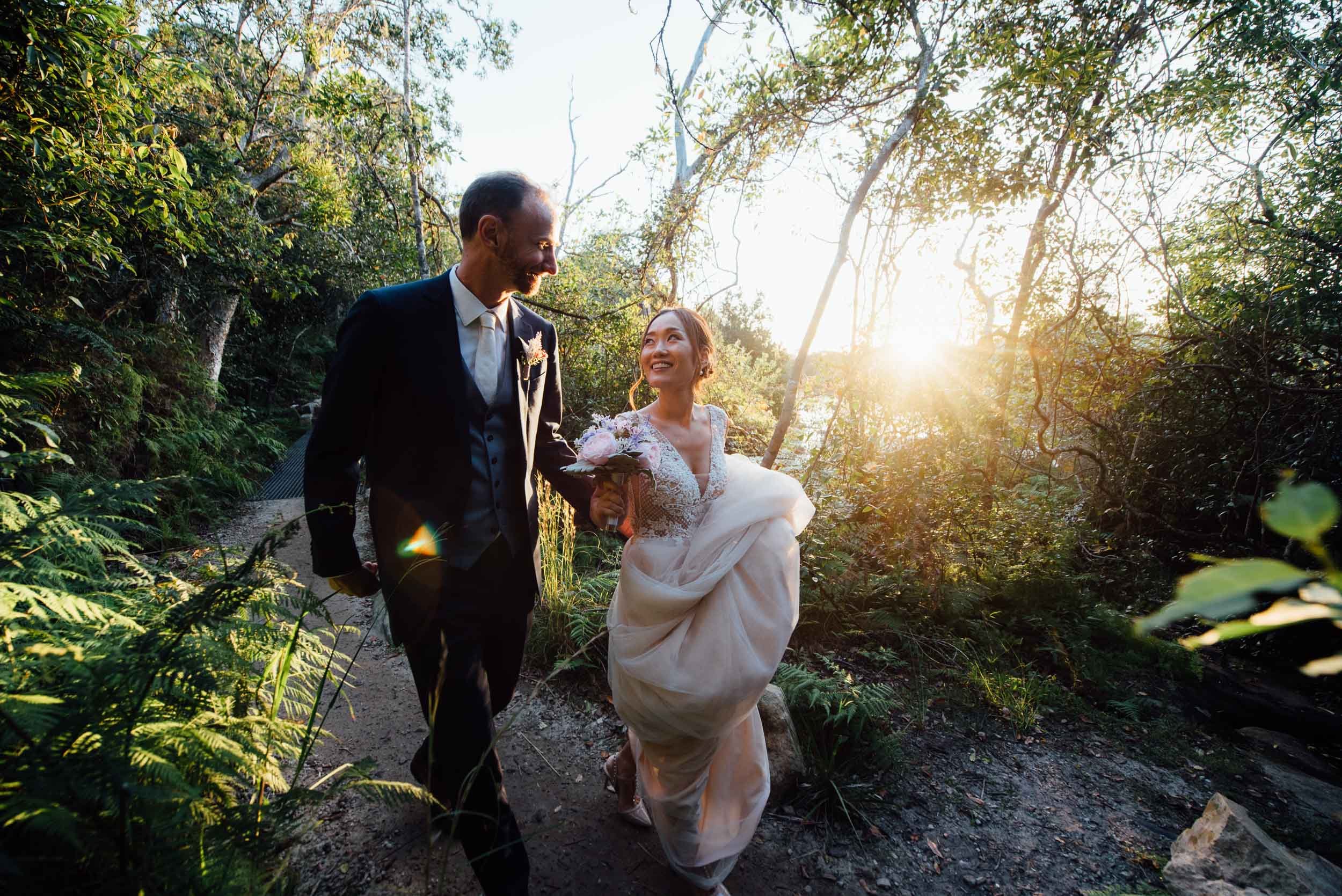 A bride and groom walking outdoors on a trail surrounded by trees, with the bride holding a bouquet, during sunset.