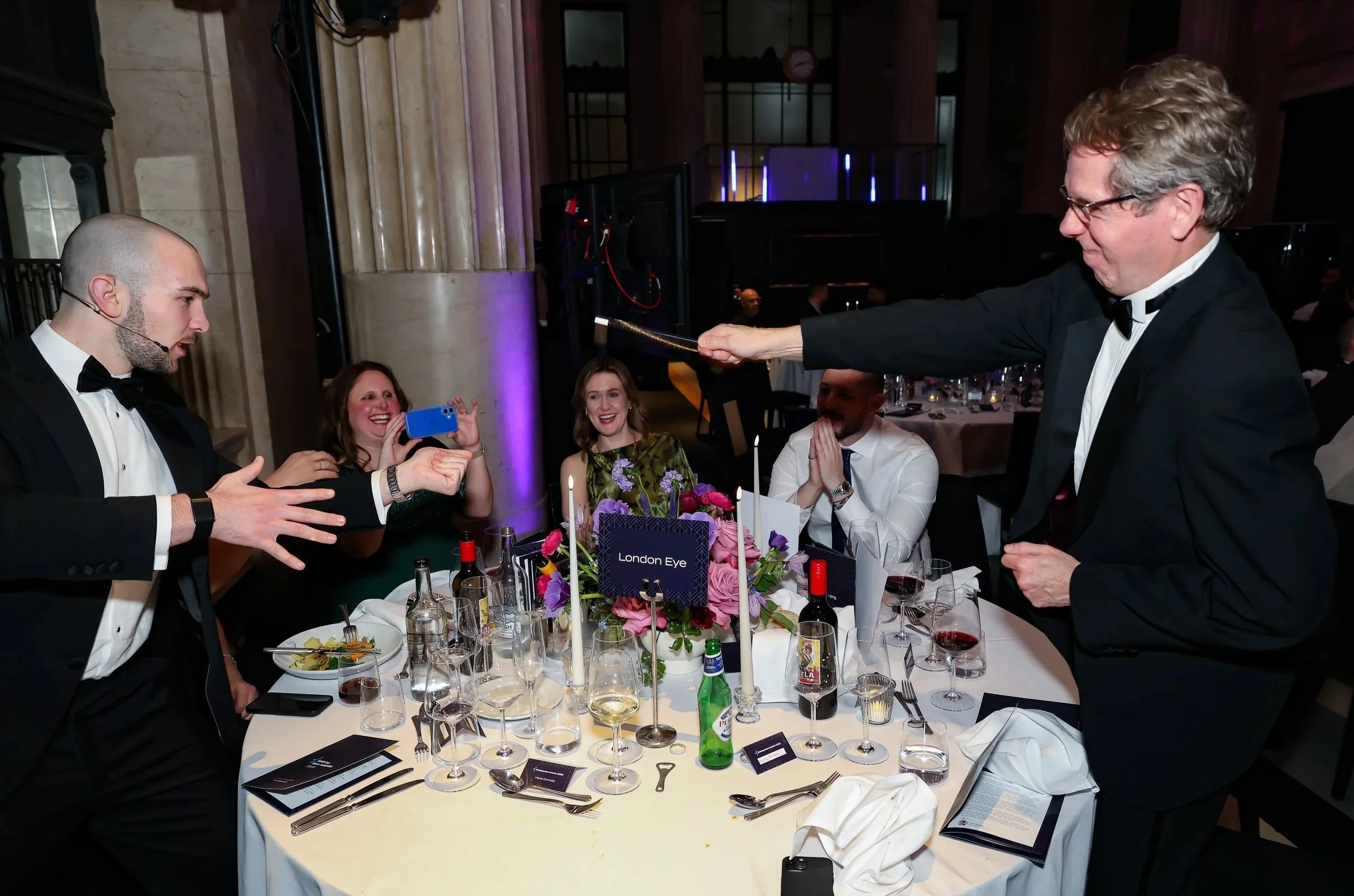A magician performing close-up magic at a corporate dinner table with guest actively participating.