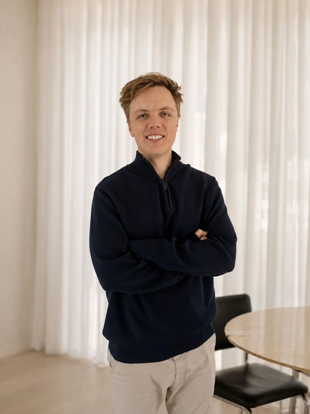 Jason Broadbridge of Buywell Group in black zip-up sweater and light-colored pants standing with arms crossed in front of sheer white curtains, in a room with a round wooden table and black chair.