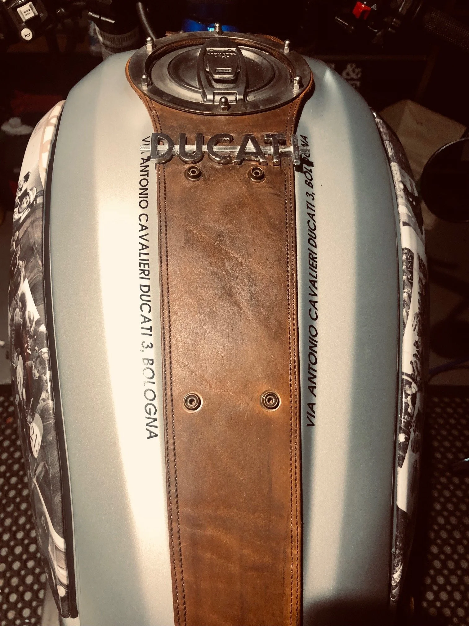 Close-up of a Ducati motorcycle fuel tank with brown leather strap and text detailing model and location.