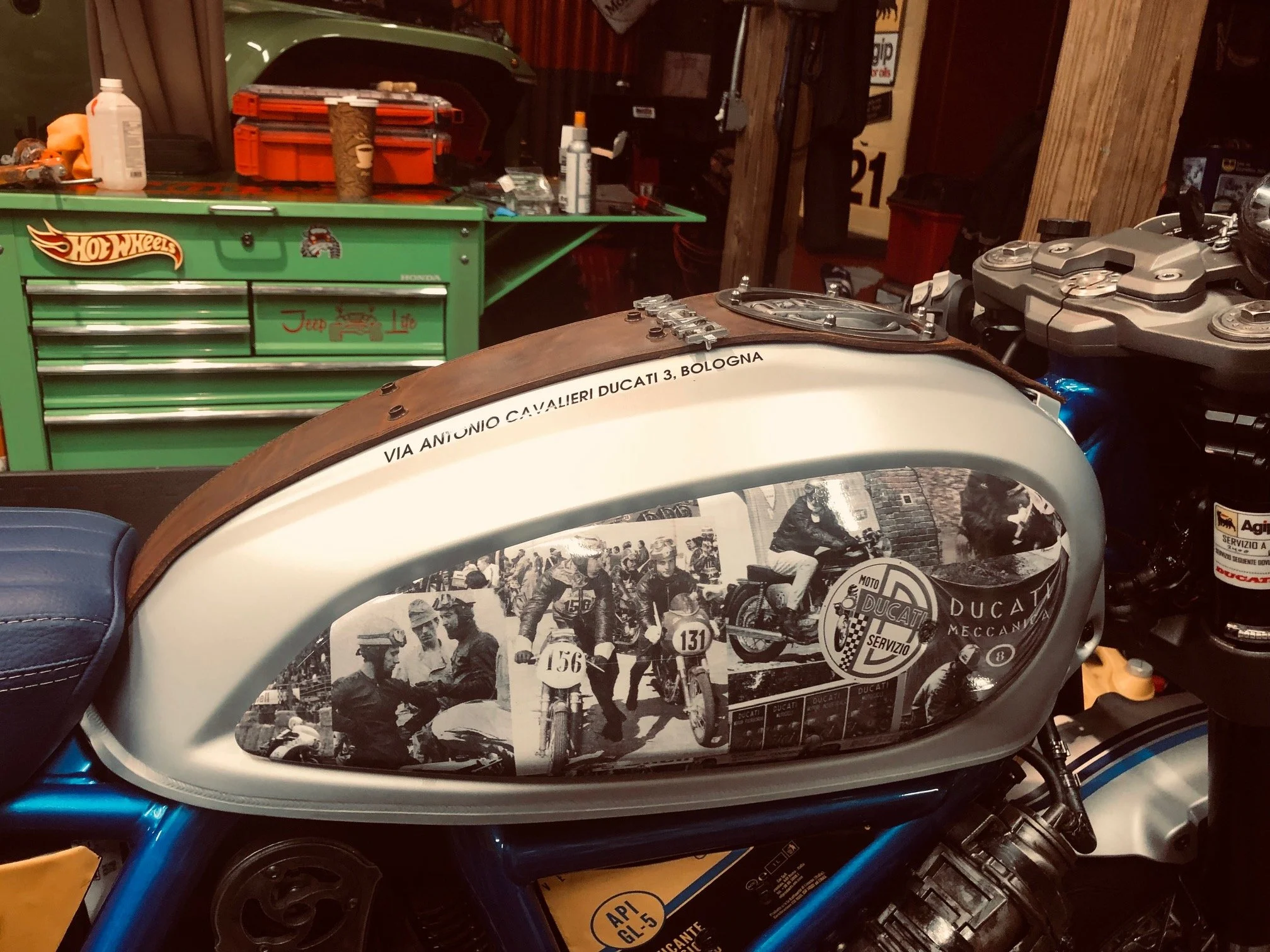 Vintage motorcycle fuel tank with Ducati branding and historical racing images in a garage with tools and green toolbox in the background.