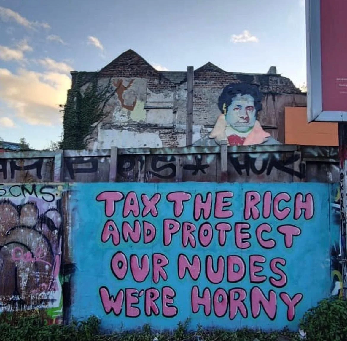 Colorful street art and graffiti on a wall, including a painting of a woman and a large sign that reads 'Tax the rich and protect our nudes were horny'.