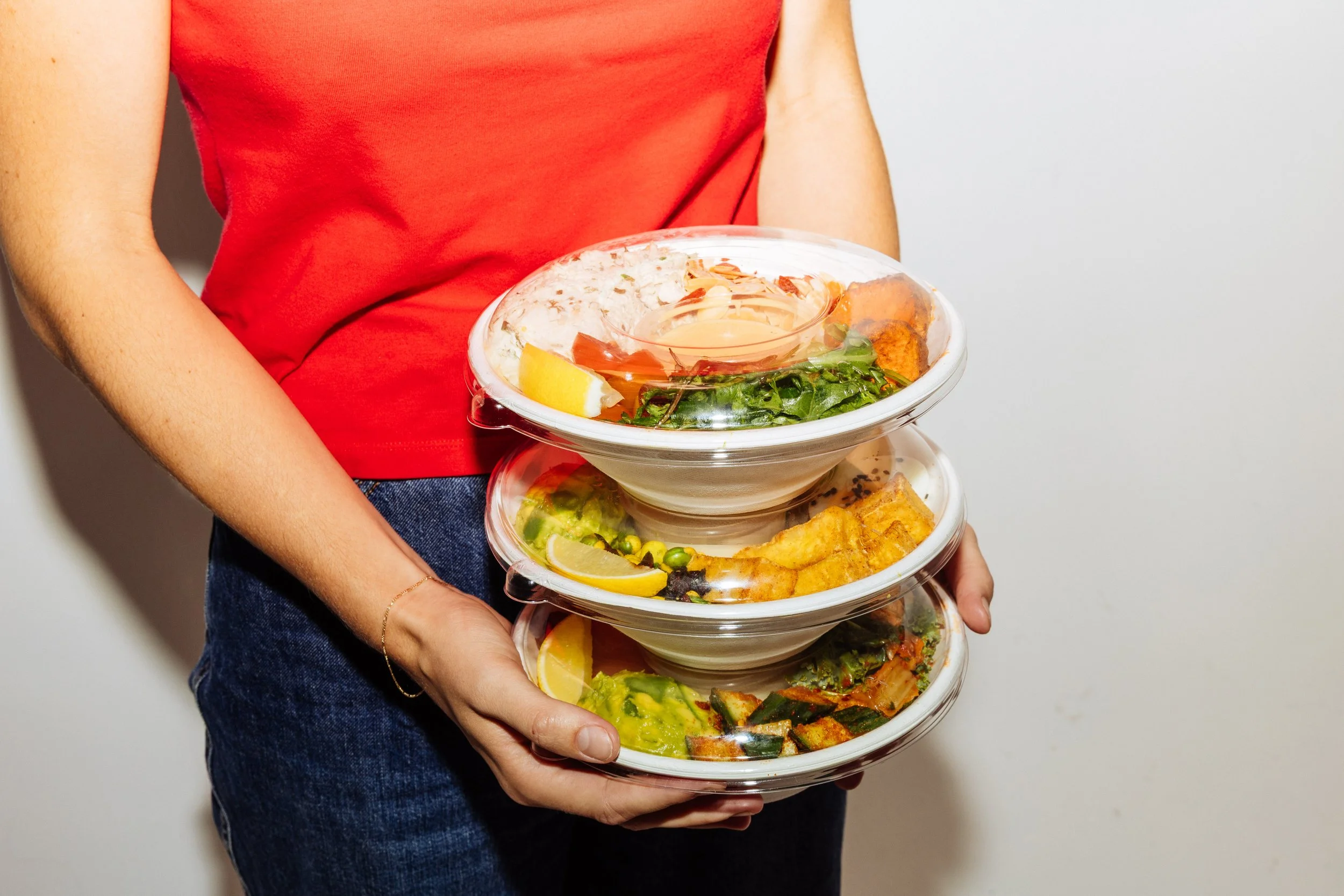 A person in a red shirt holding a stack of three transparent food containers filled with salads and side dishes, with a plain wall in the background.