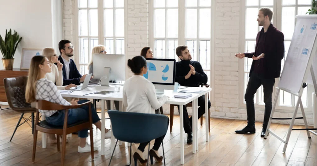 A business presentation in a modern, well-lit office with seven people and a man standing at a whiteboard.