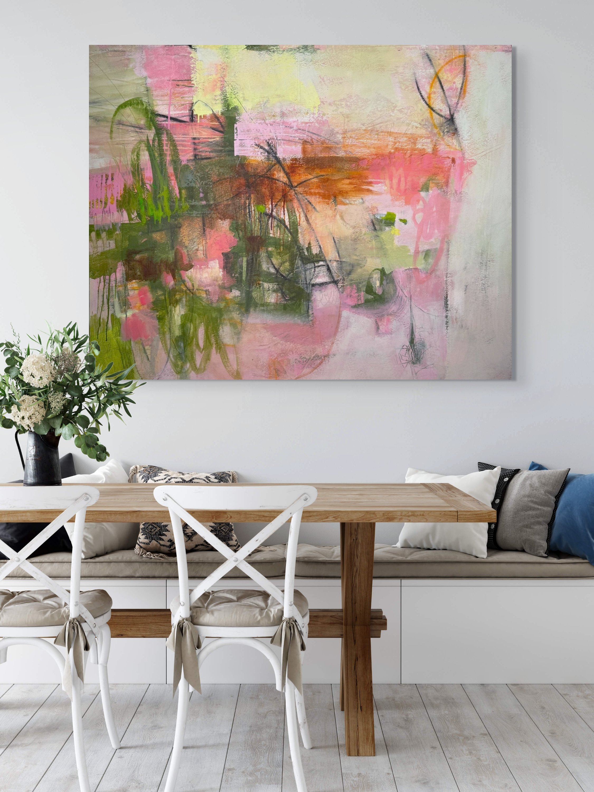 A dining area with a wooden table, white chairs with beige ties, a beige cushioned bench with pillows, a floral bouquet on the table, and an abstract pink, orange, and green painting on the white wall.