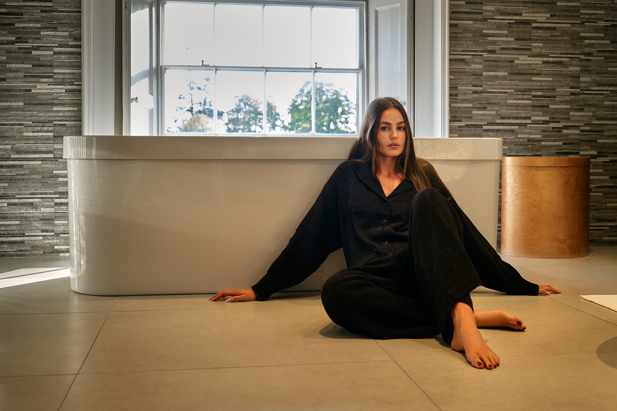 Woman in black pajamas sitting on the floor next to a bathtub near a window with trees outside.