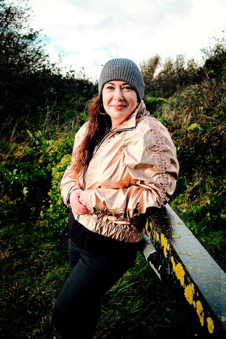 A woman outdoors leaning on a yellow and black painted railing, wearing a gray knit beanie and a shiny rose gold jacket.