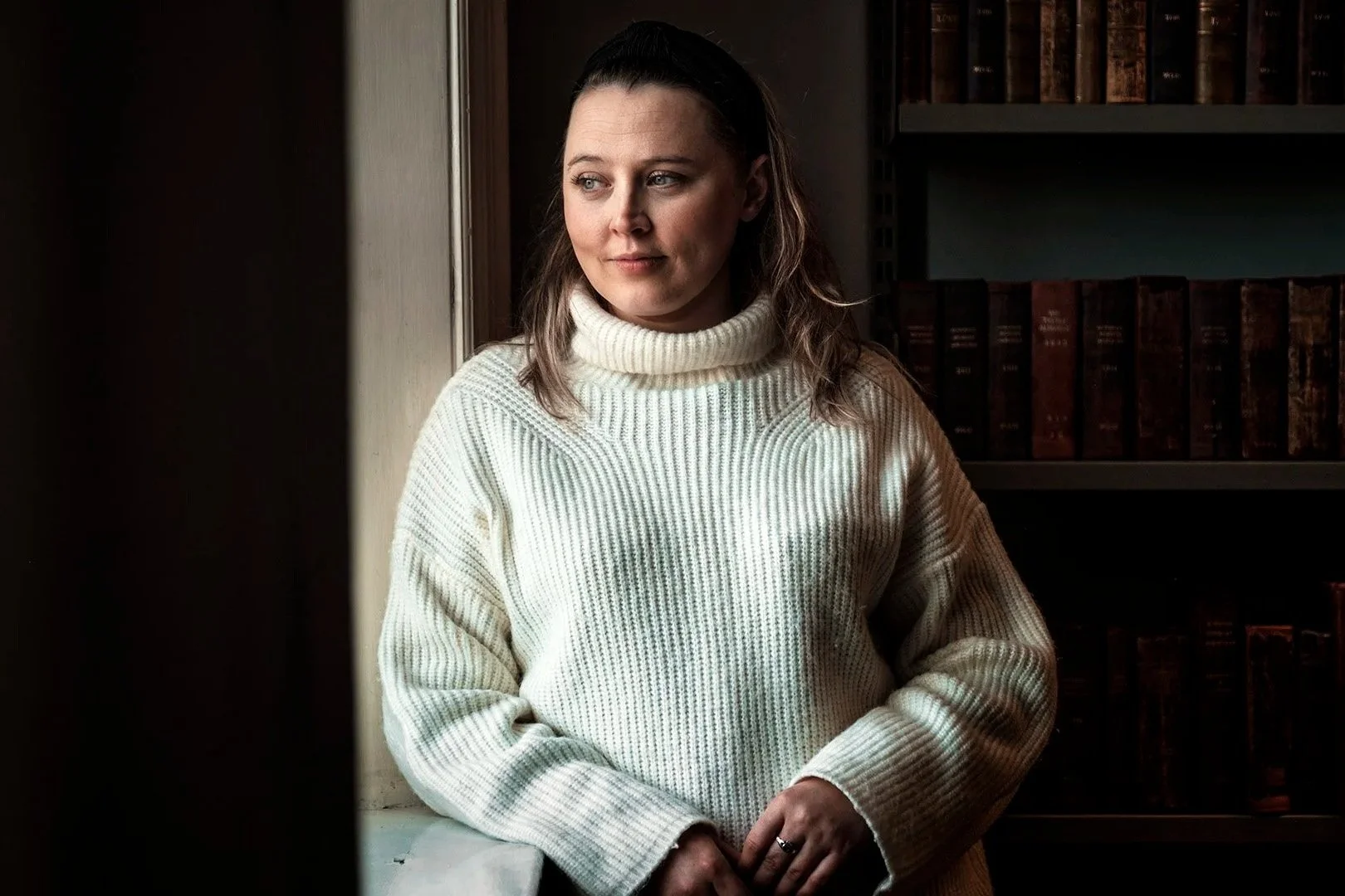 Woman in white sweater leaning against window with bookshelves in the background.