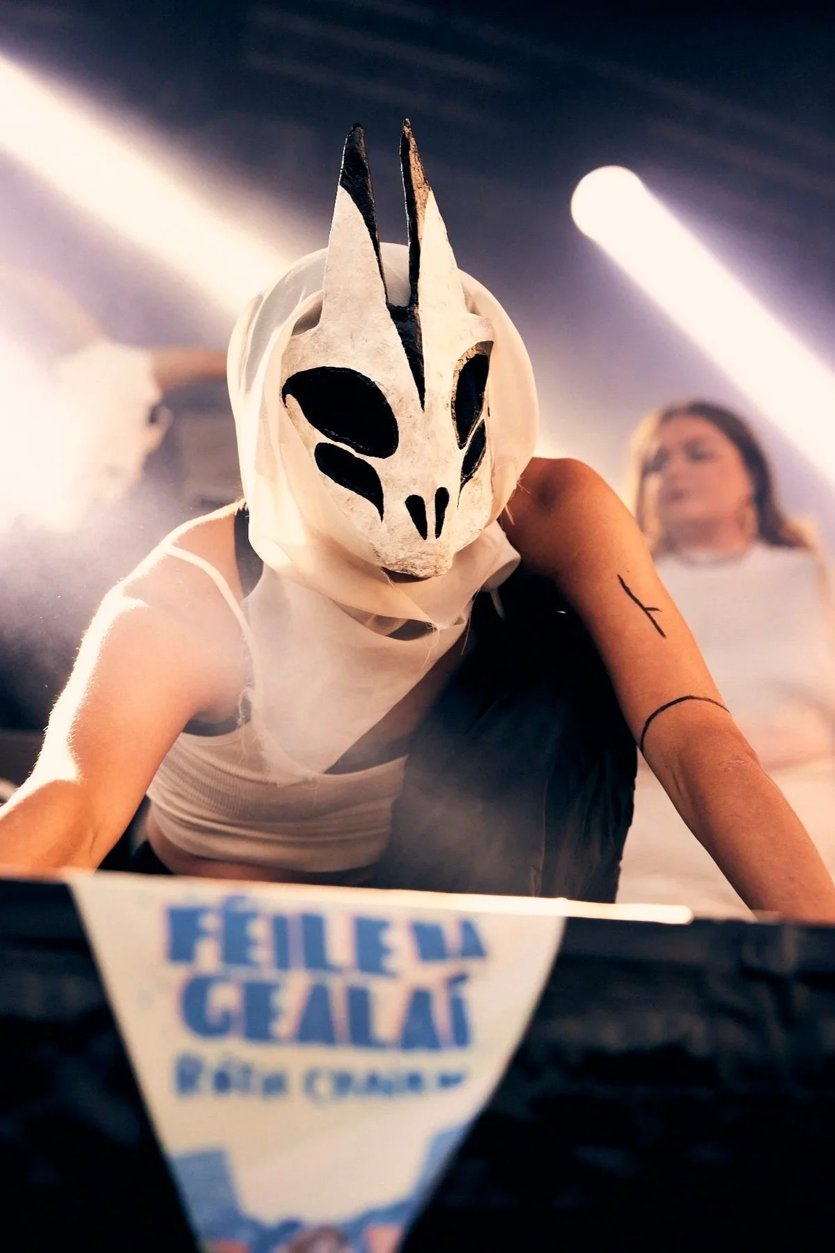 Person wearing a white and black mask with animal-like features, performing on stage with festival banner in foreground and spotlight in background.