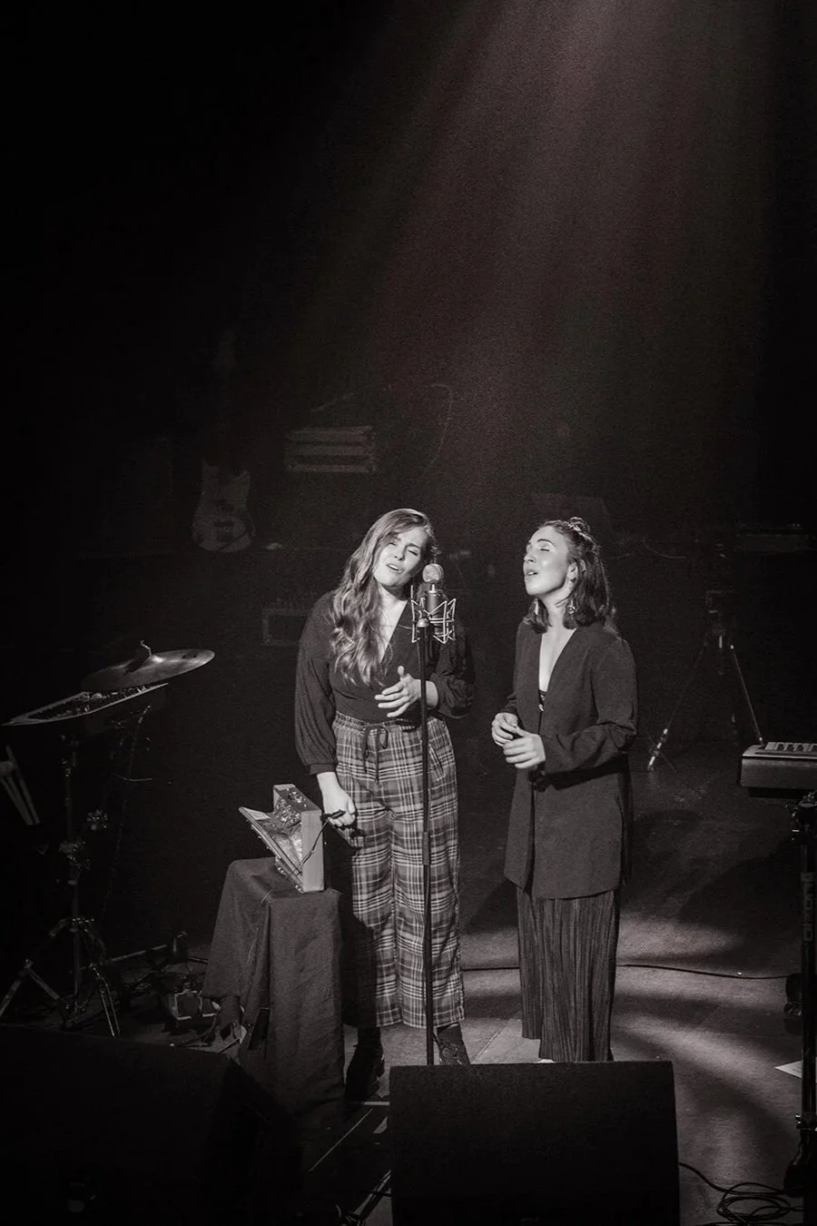 Two women singing on stage in a spotlight, with music equipment around them. One is holding a microphone stand and both are wearing dark outfits. The setting is dimly lit, creating a dramatic atmosphere for the performance.
