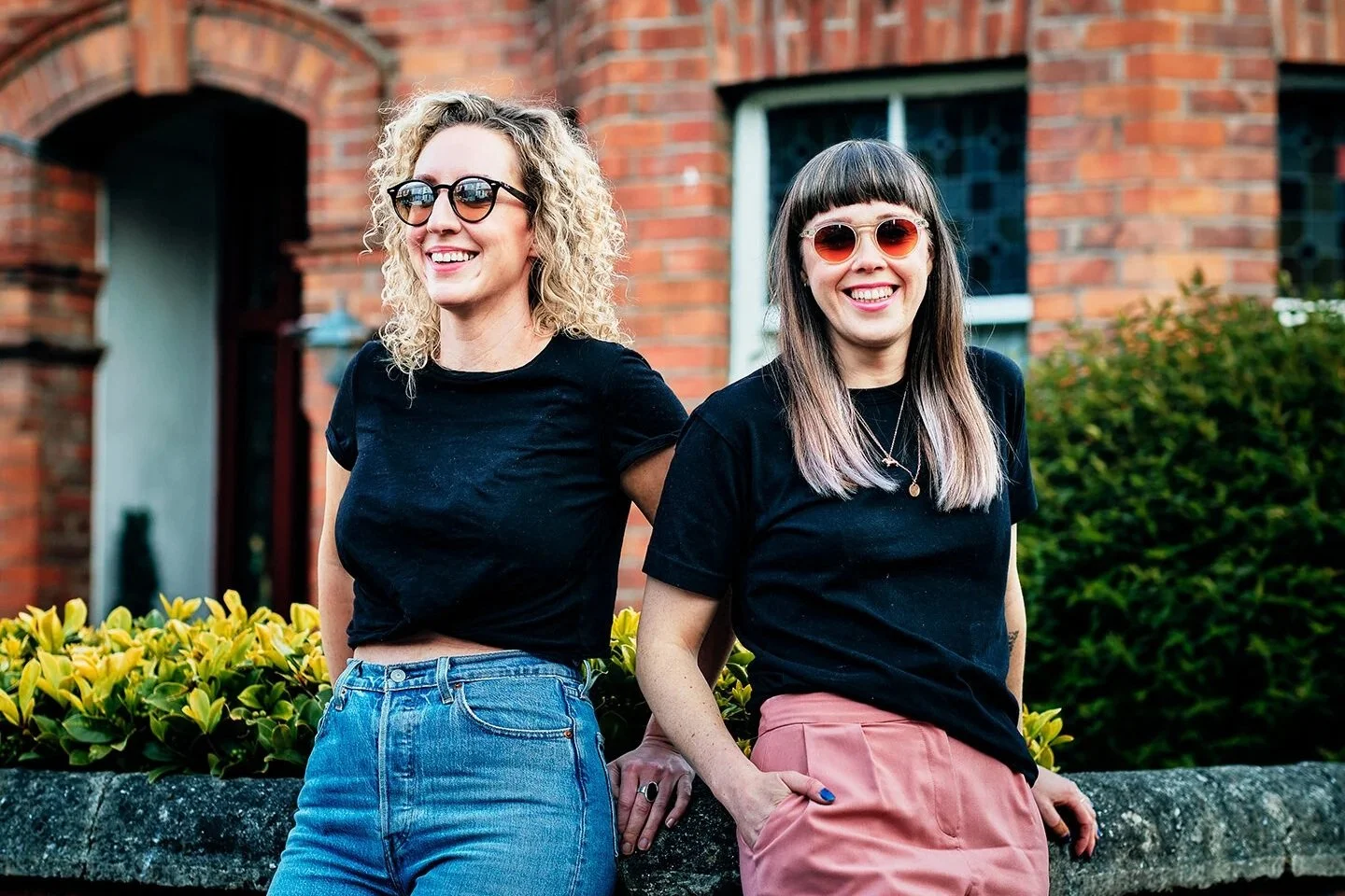 Two smiling women standing outdoors in front of a brick house and green bushes, wearing black t-shirts and sunglasses.