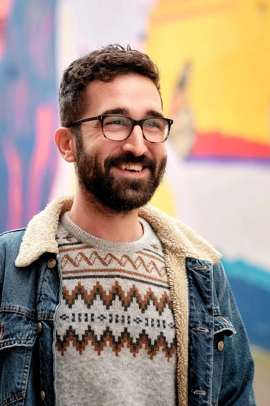 A smiling man with glasses, a beard, and short brown hair, wearing a patterned sweater and a denim jacket with a shearling collar, standing in front of a colorful abstract background.