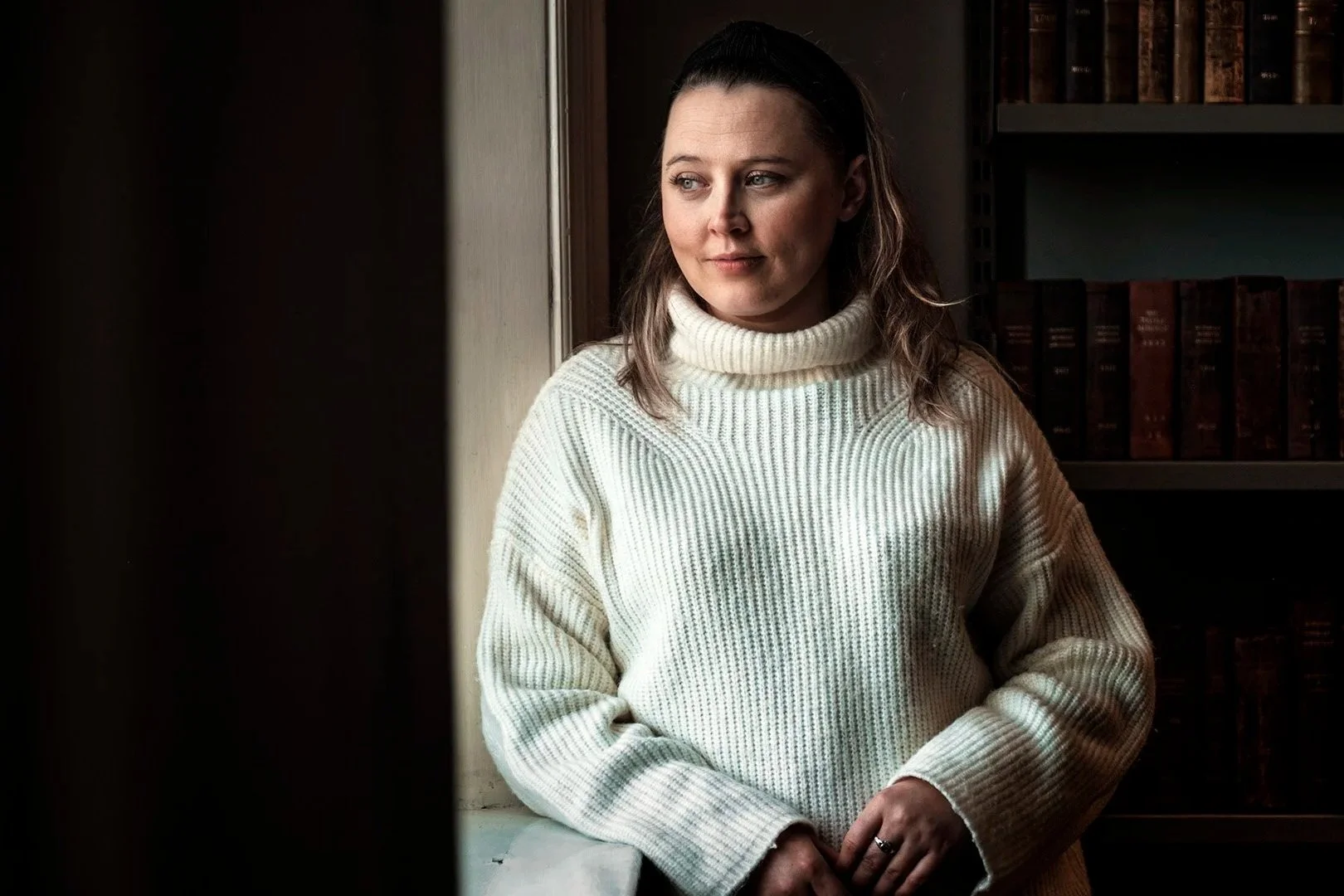 Person in white turtleneck sweater standing by window with bookshelf in background.
