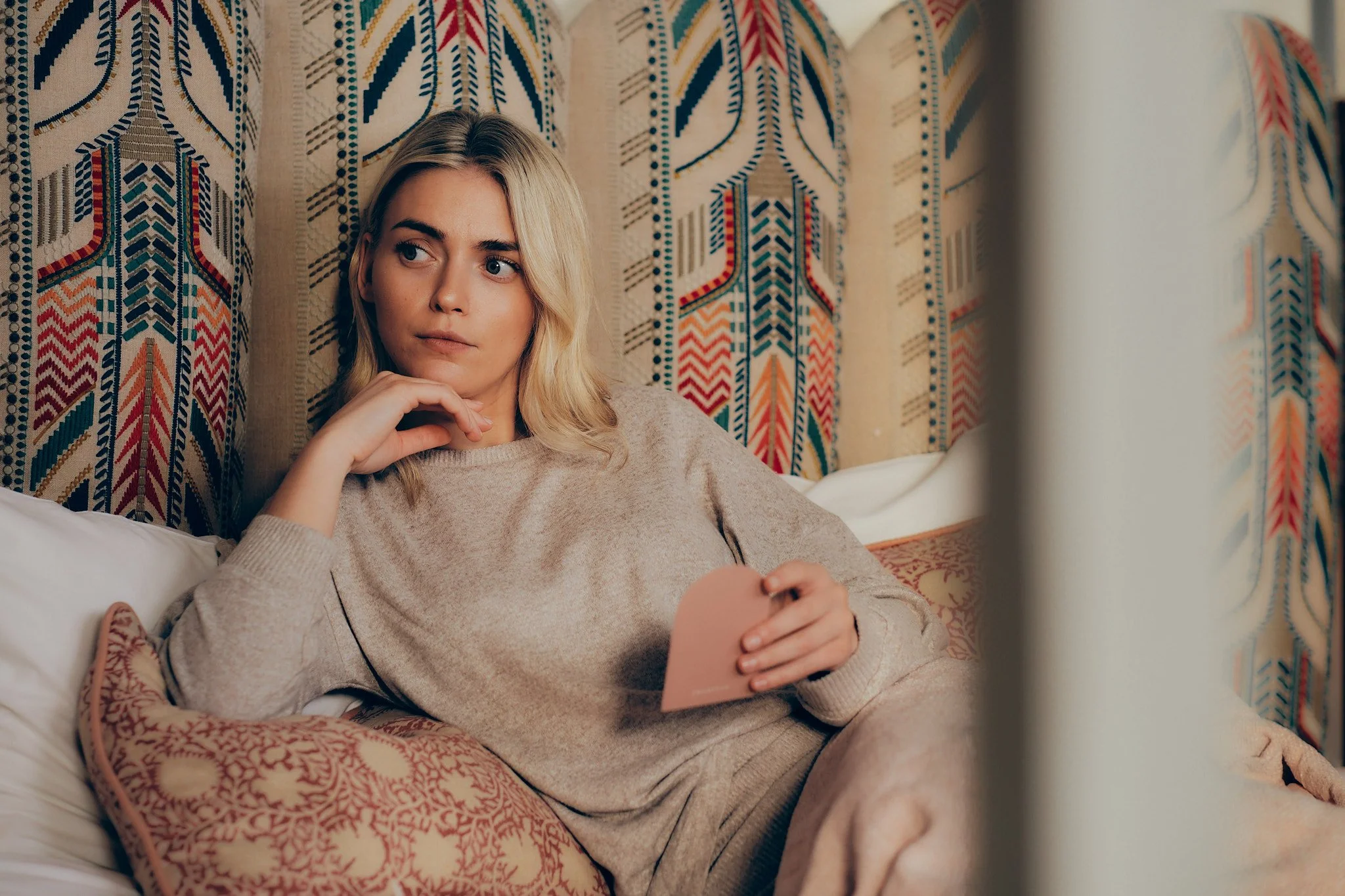 A young woman with blonde hair lying on a bed, resting her head on a pillow, holding a pink mirror, with patterned curtains behind her.