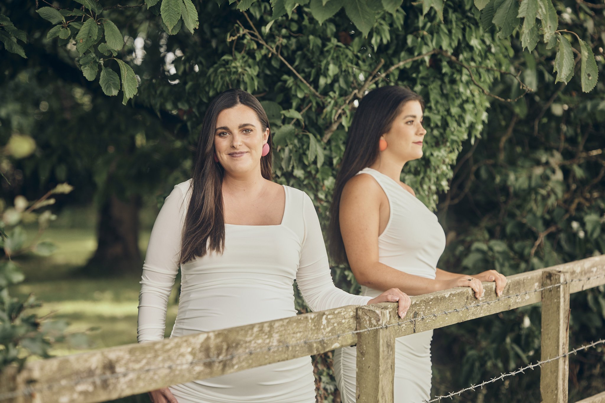 Two women standing by a rustic wooden fence outdoors with green foliage in the background, one woman facing forward and the other in profile, both wearing white dresses and colorful earrings.